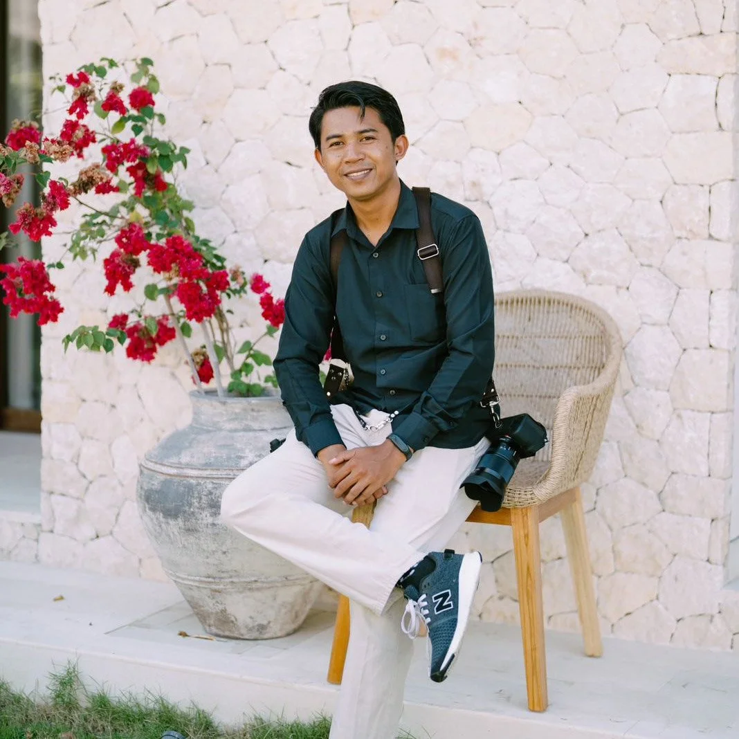 A man wearing a dark teal shirt and white pants sitting on a small wooden stool, smiling at the camera. He has a camera hanging from his shoulder and a walkie-talkie clipped to his shirt. Behind him is a large potted plant with red flowers and a stone wall.