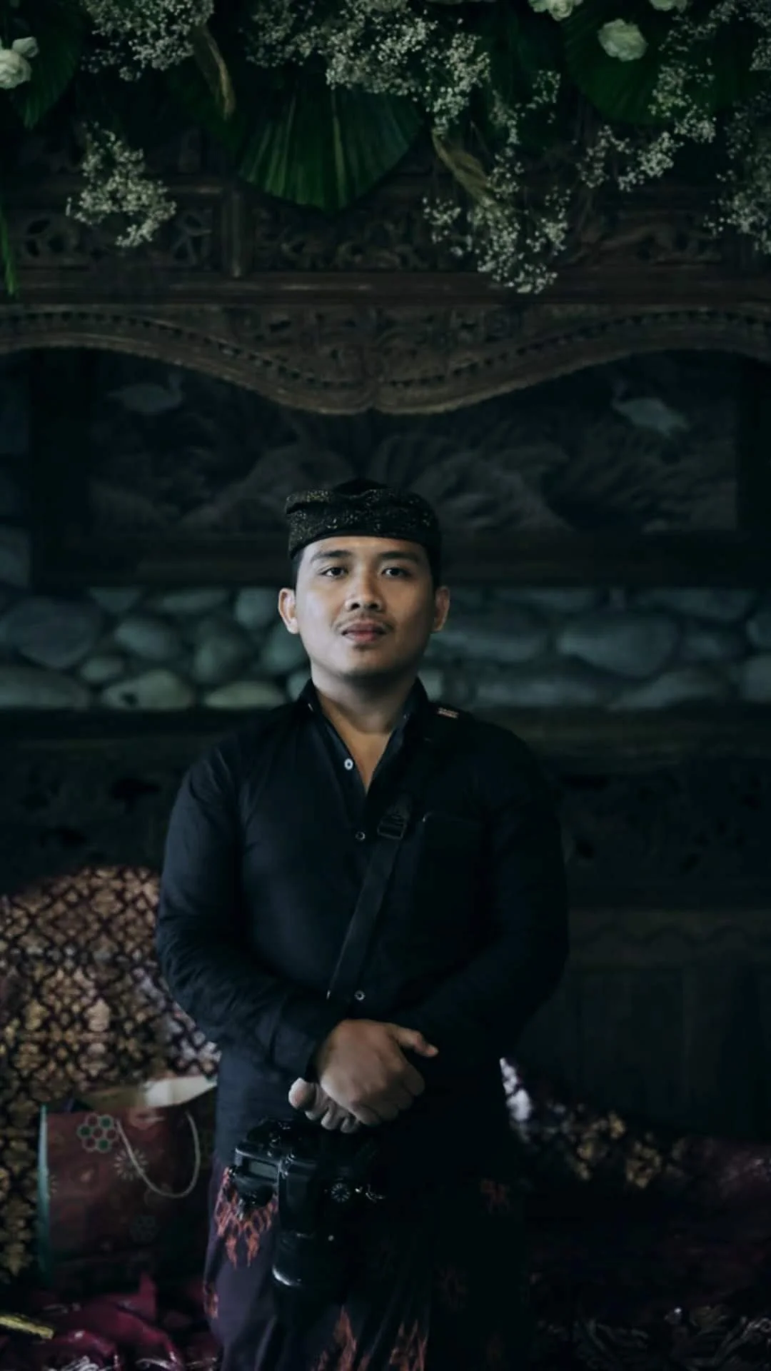 A man in traditional Indonesian attire stands indoors, holding a camera. The background features wooden carvings, stonework, and floral decorations.