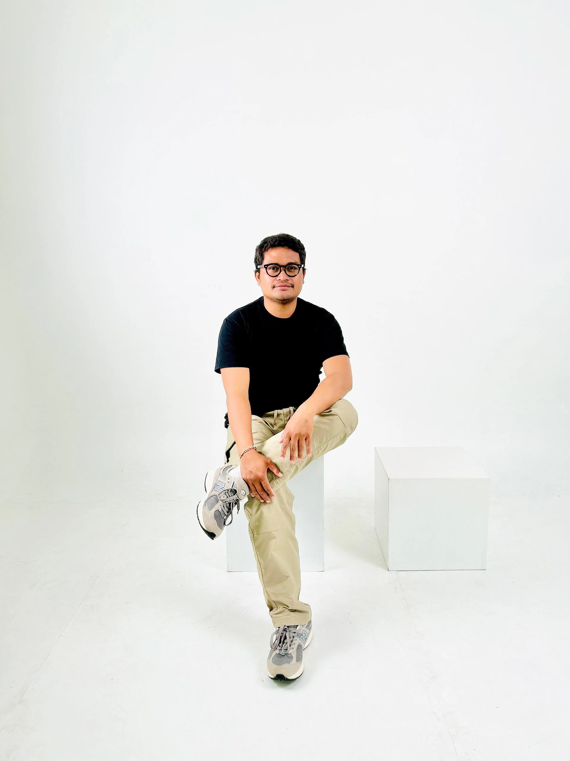 A man with glasses wearing a black t-shirt and khaki pants, sitting on a white block in a white studio, with another white block nearby.