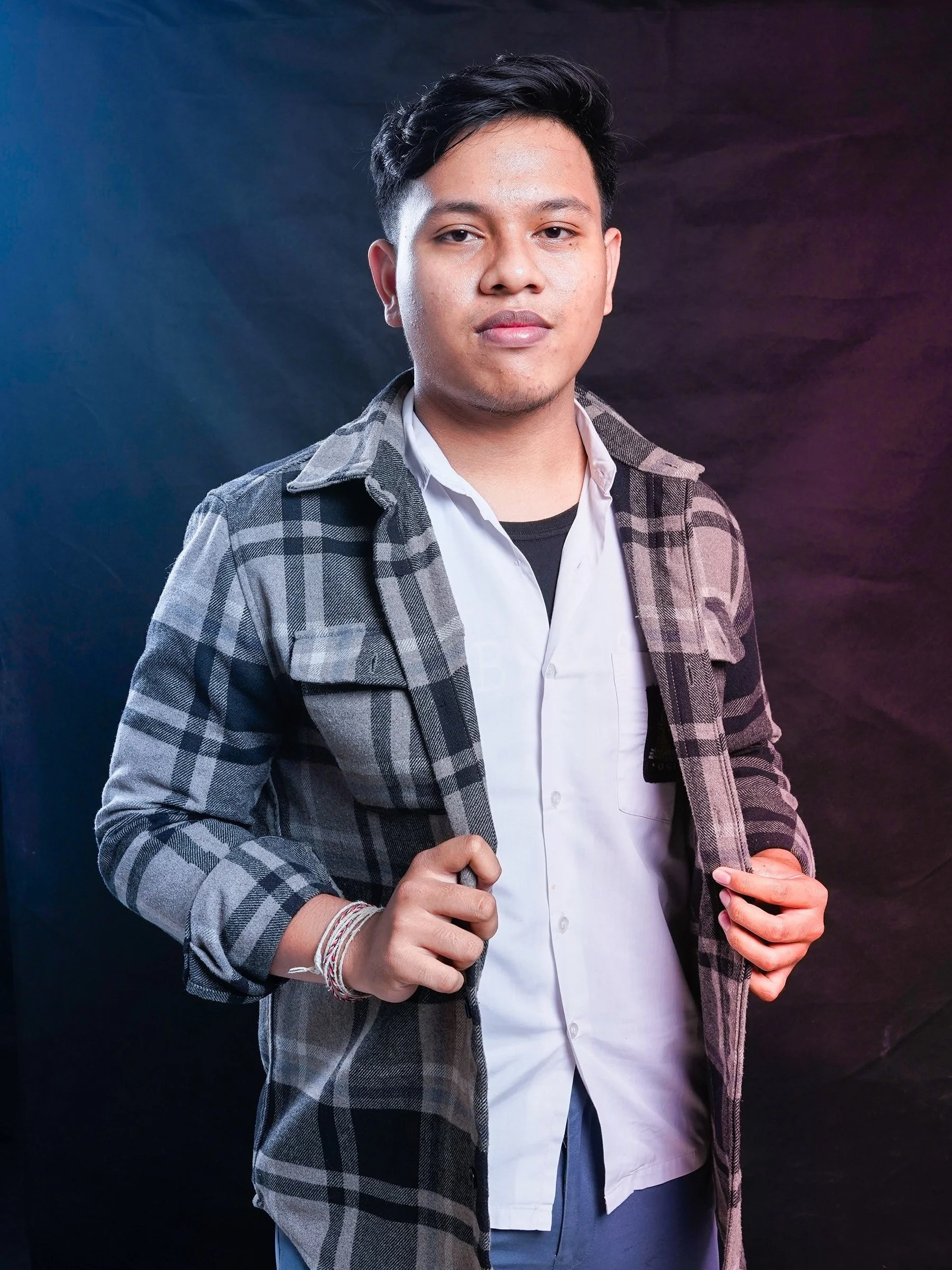 A young man with dark hair, wearing a white button-up shirt layered over a black shirt, and a gray plaid jacket, standing in front of a dark background with colorful lighting.