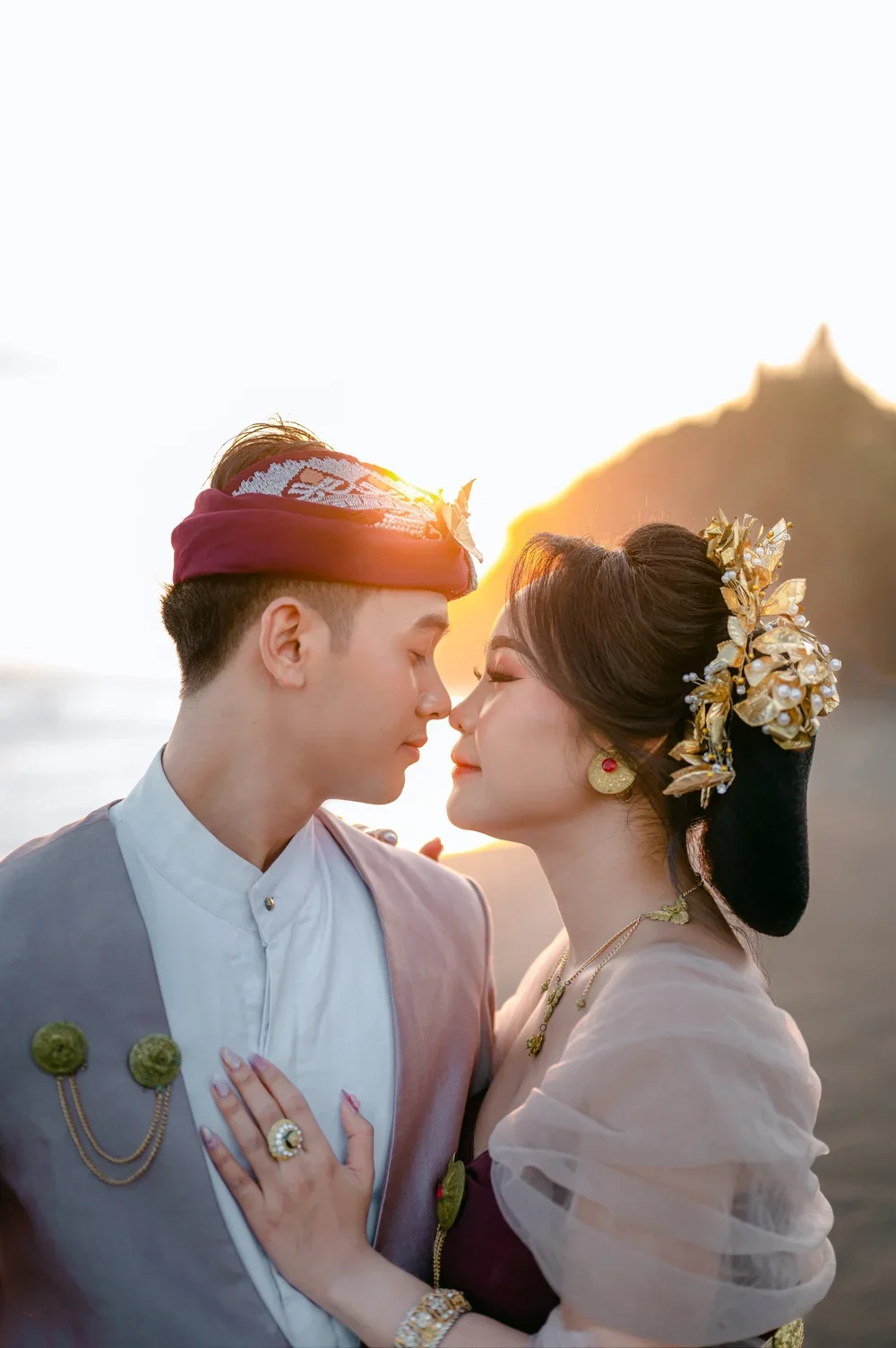 An Asian couple dressed in traditional attire, standing close with noses touching on a beach at sunset, with a mountain in the background.