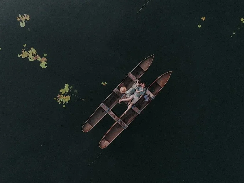 Two children lying on a wooden raft in a dark, calm body of water with some floating lily pads nearby.