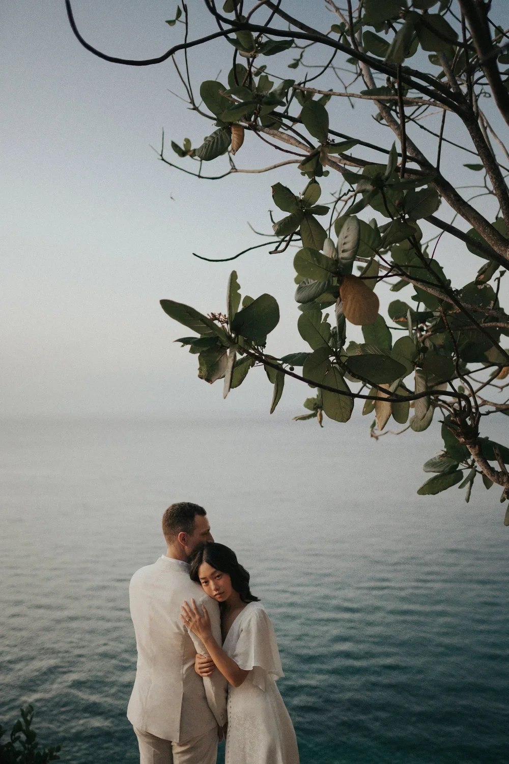 A couple in white clothing embrace near a body of water with a tree and foliage in the foreground, during dusk or early evening.
