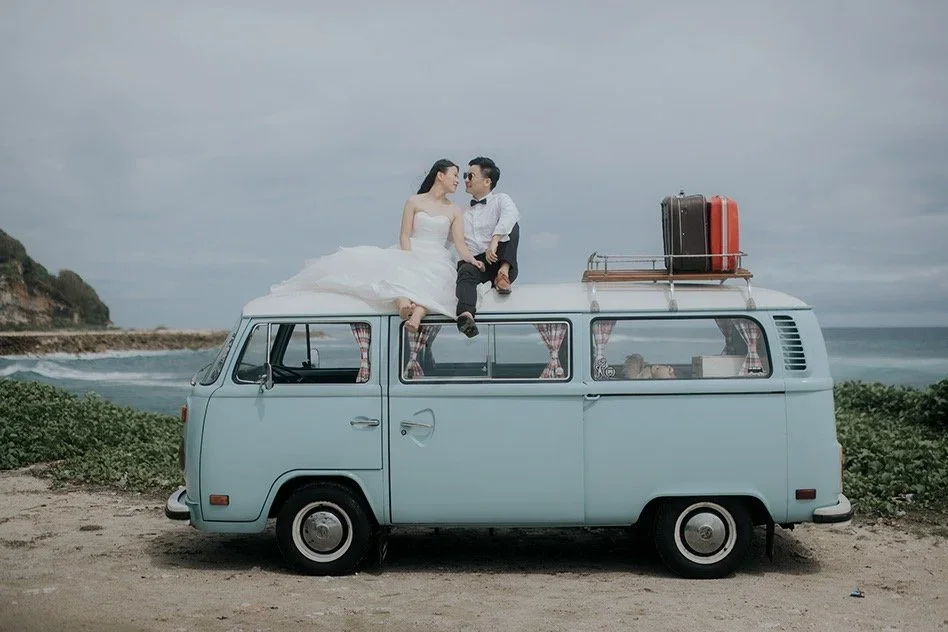 A couple dressed in wedding attire sitting on the roof of a vintage light blue camper van on a beach, with suitcases and the ocean in the background.