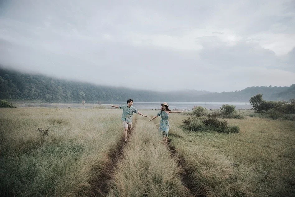 A couple holding hands and walking on a dirt path through a grassy field near a body of water with distant mountains, overcast sky.