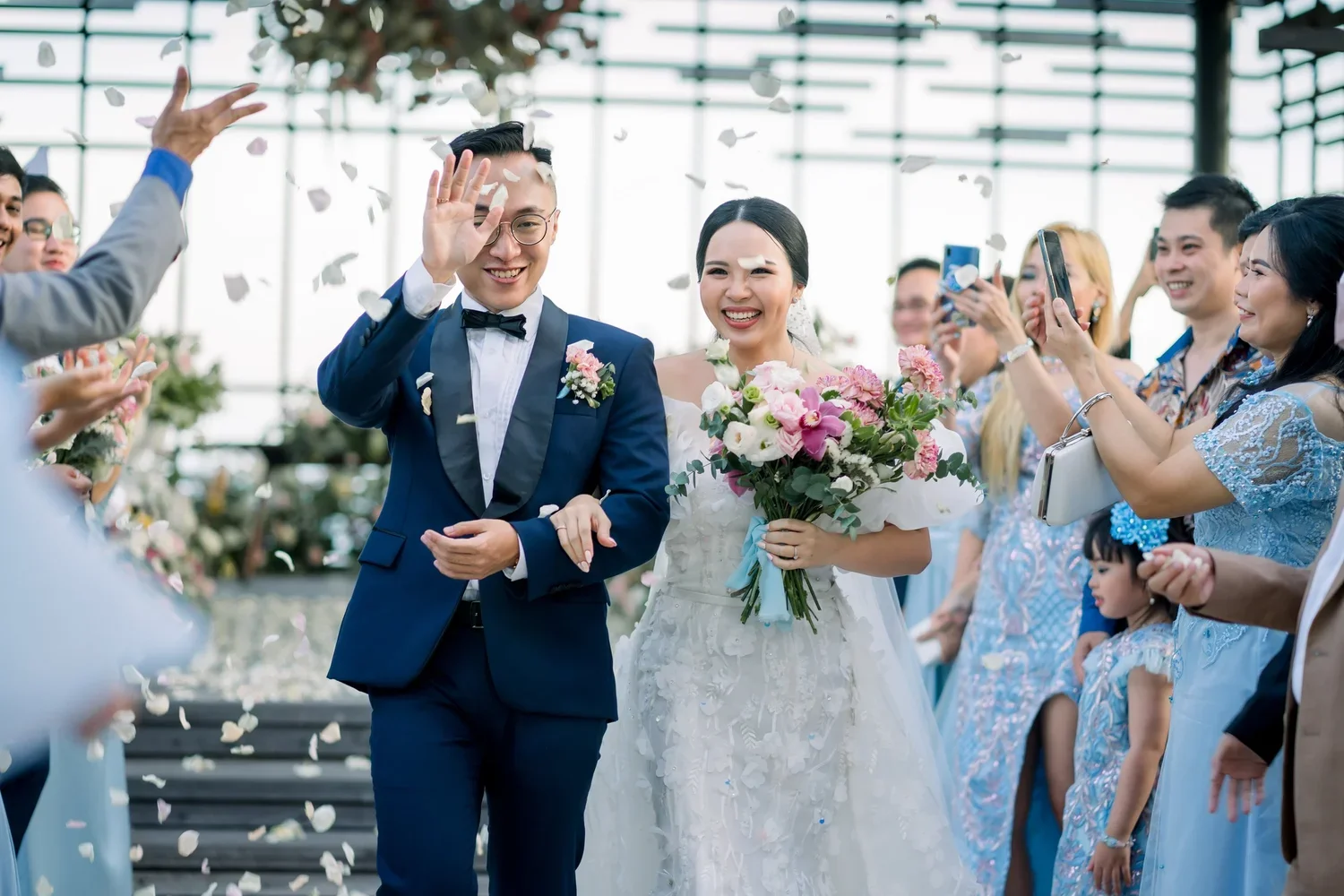 A newlywed couple smiling and celebrating as friends and family throw flower petals at them during a wedding ceremony.
