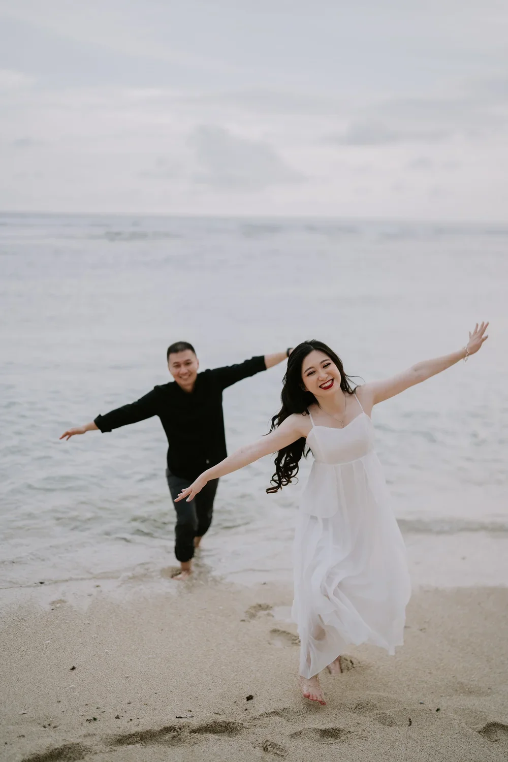 A smiling woman in a white dress and a man in black shirt and pants are playing and running on the beach near the water, with arms outstretched and joyful expressions.