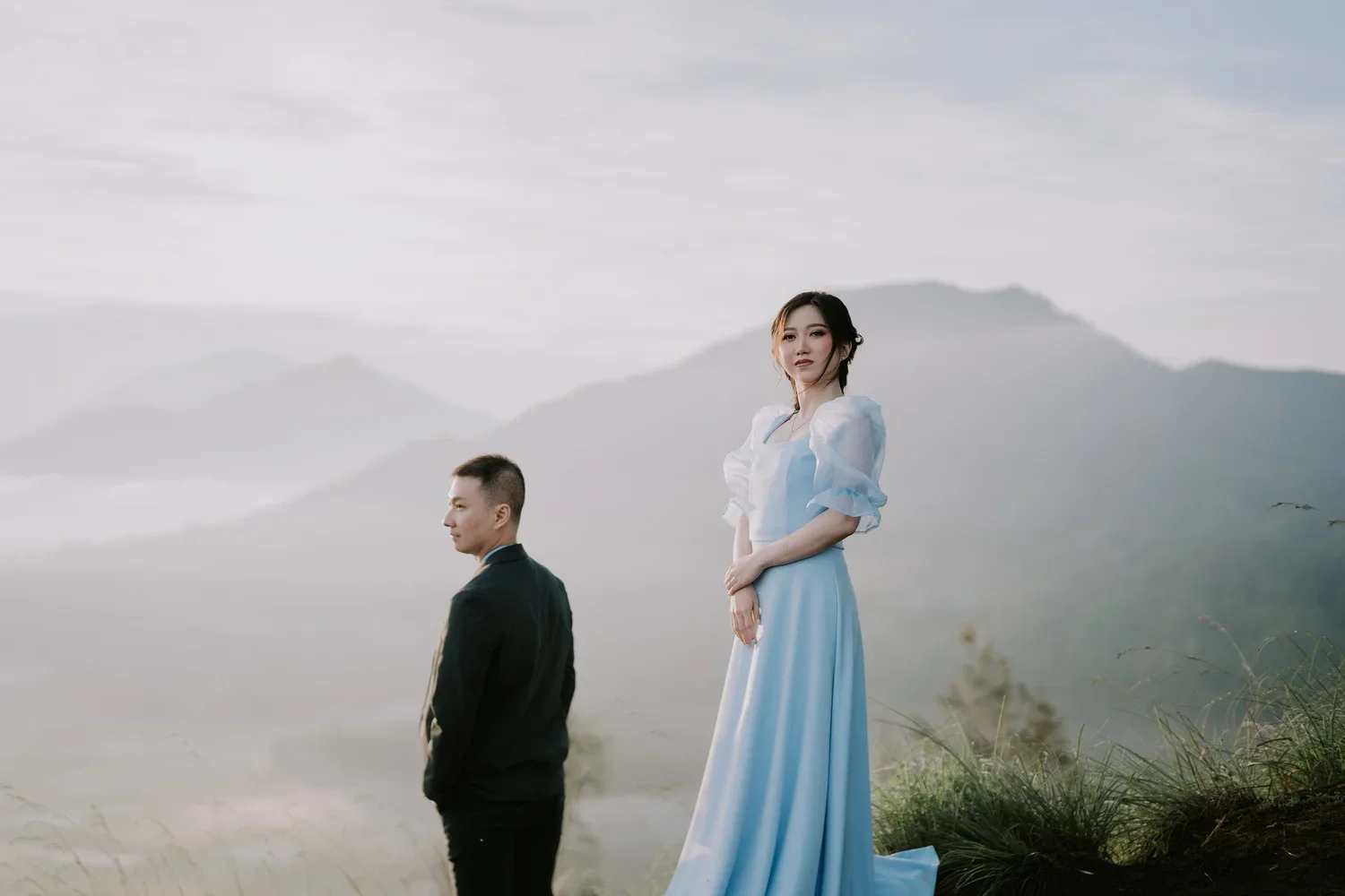 A man in a black suit and a woman in a blue dress standing outdoors on a grassy hill with mountains and a cloudy sky in the background.