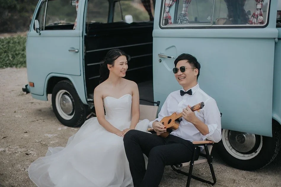 A woman in a white wedding dress and a man in sunglasses wearing a white shirt with a black bow tie, playing a ukulele, sitting in front of a vintage light blue van.