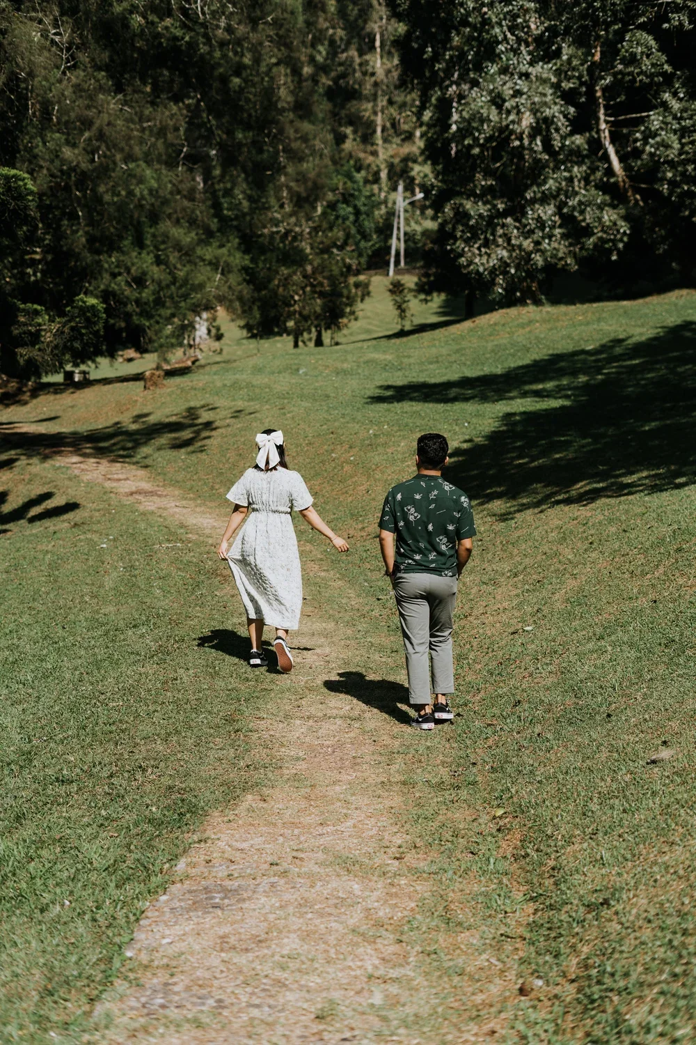 A woman in a white dress walking on a dirt path with a man in a dark shirt and light pants in a green park with trees around.