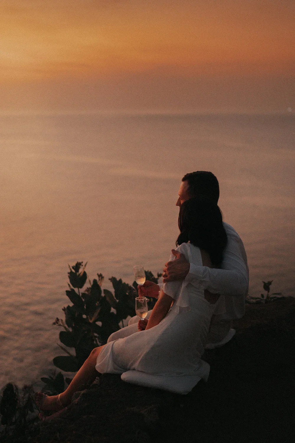 A couple sits on a cliffside during sunset, embracing and holding glasses of champagne with ocean and colorful sky in the background.