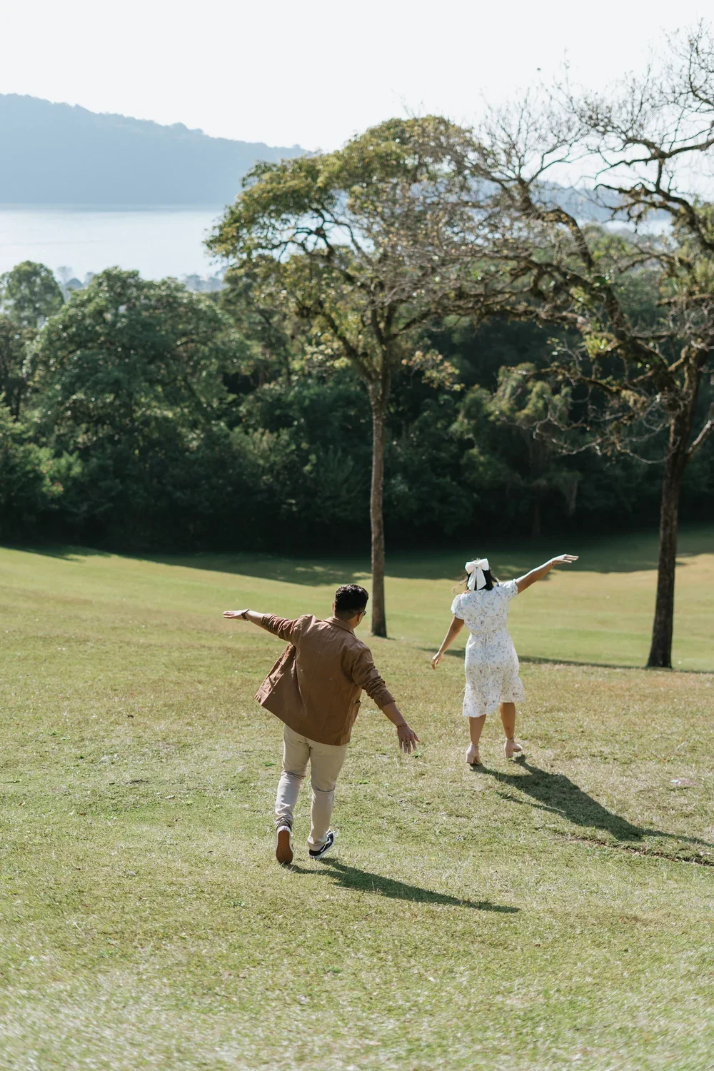 A couple running and playing in a grassy park with trees and mountains in the background.