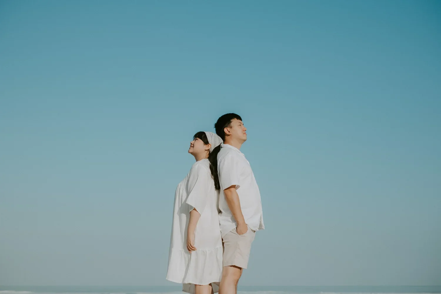 A man and woman stand back-to-back in a beach setting with clear blue sky, both with eyes closed and smiling softly, dressed in white clothing.
