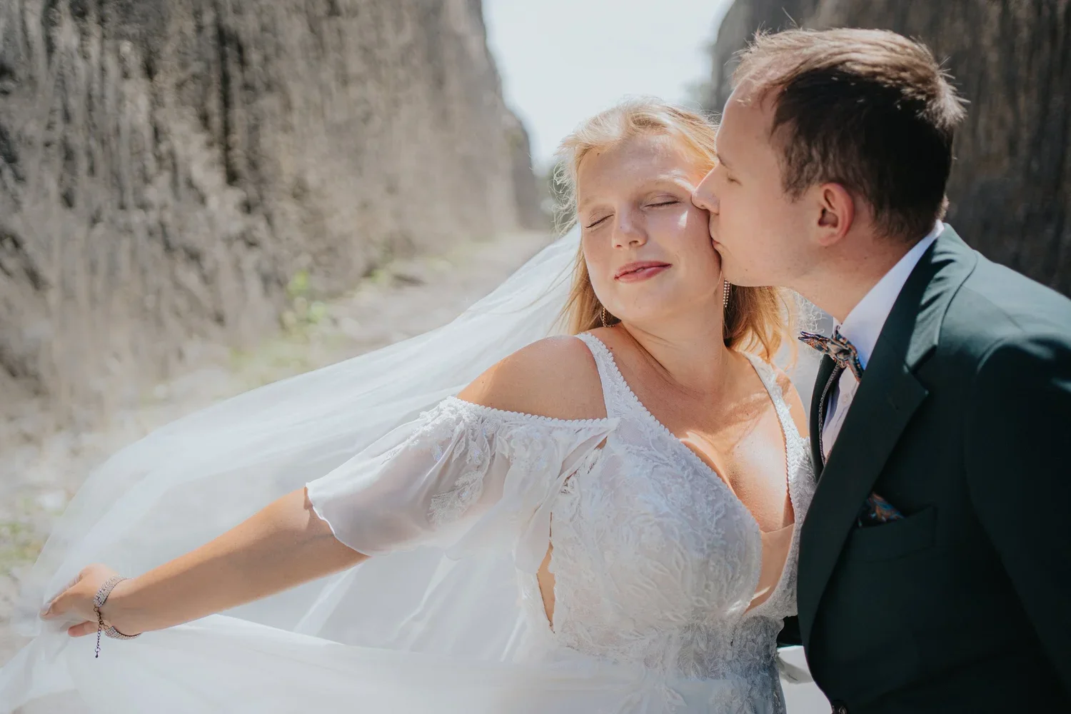 A bride in a white wedding dress and a groom in a dark suit share an intimate moment outdoors, with the groom kissing the bride on the cheek as she closes her eyes and smiles.
