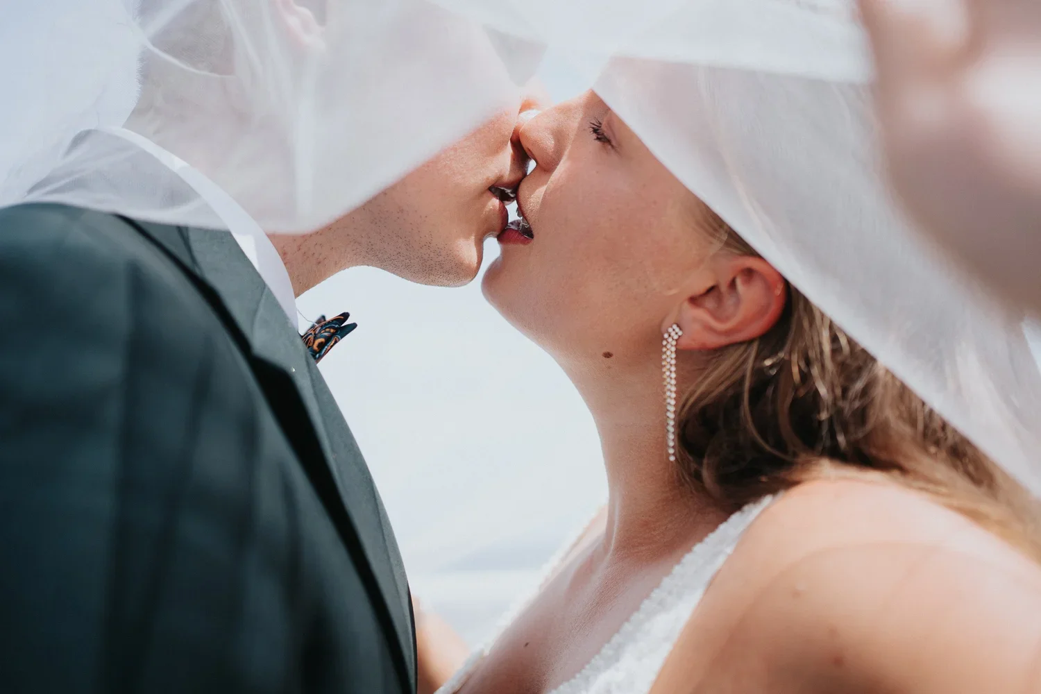 Close-up of a bride and groom sharing a kiss under a white veil, with the bride wearing an earring and the groom in a dark suit.