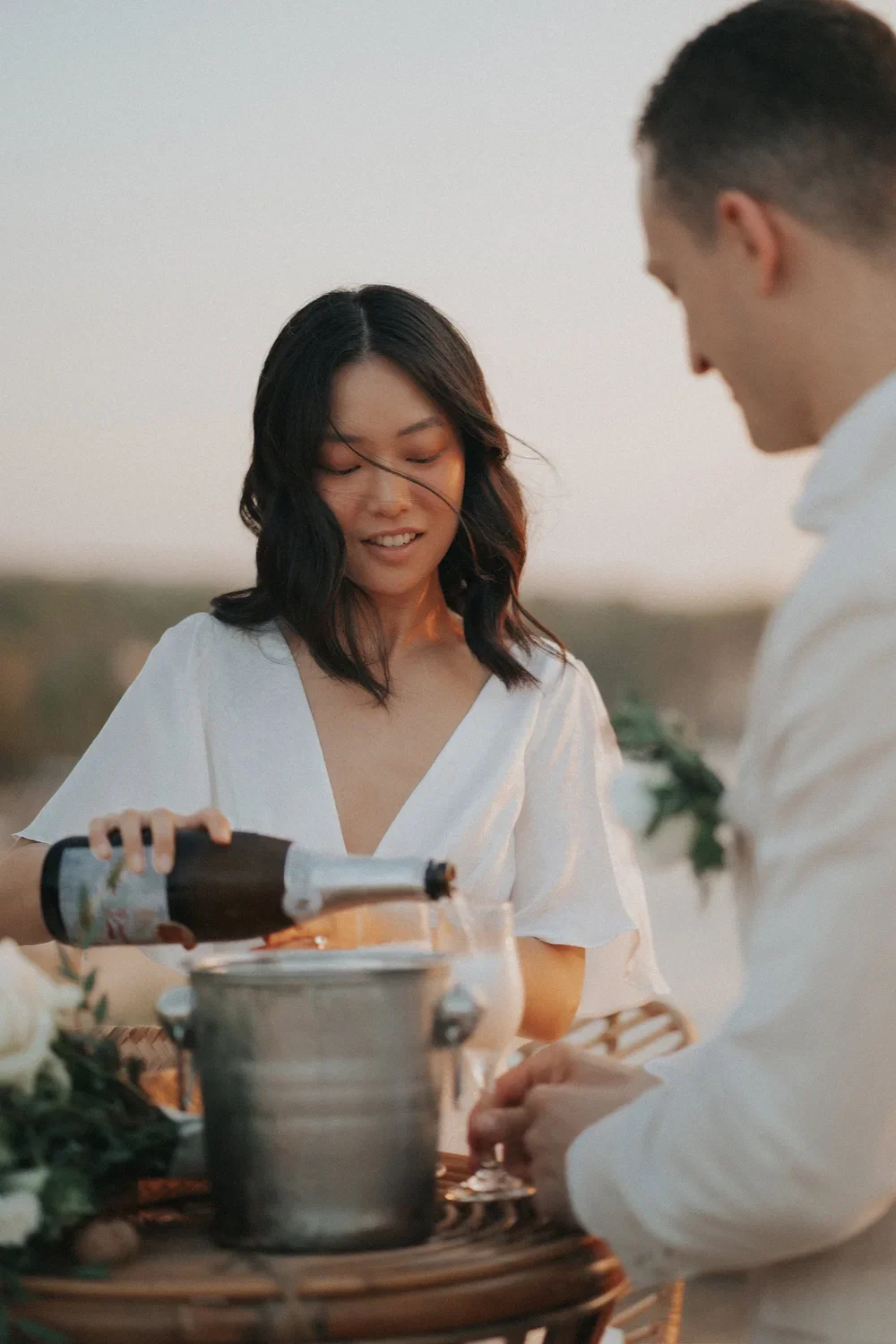 A woman pours champagne into a glass during an outdoor celebration at sunset, with a man standing beside her.