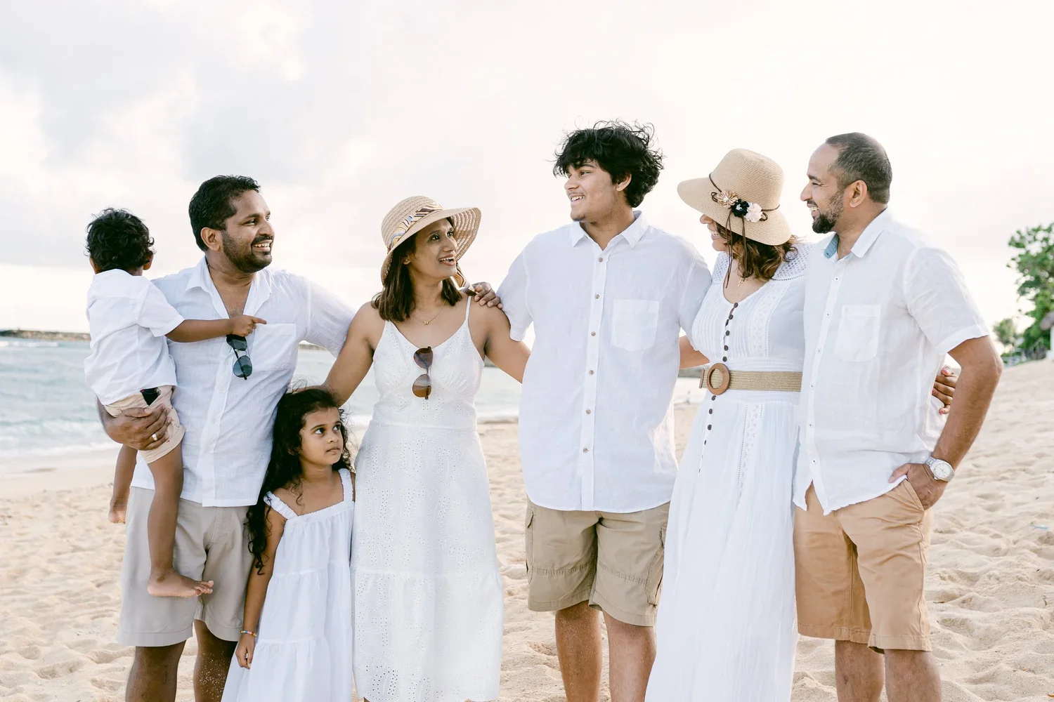 A group of seven people on the beach, smiling and talking, with two children, wearing summer clothes and hats, enjoying a sunny day by the ocean.