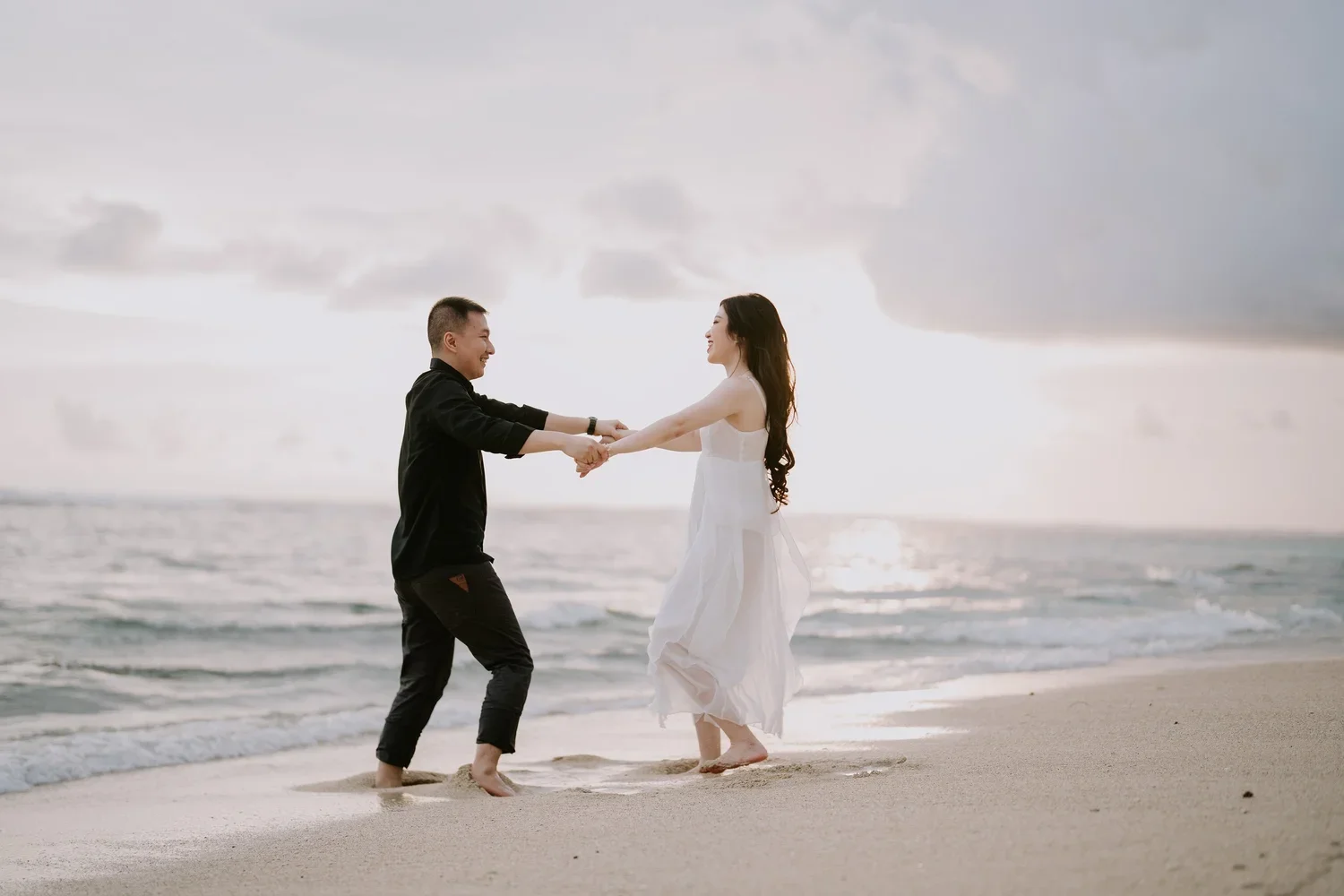 A couple holding hands and smiling on the beach at sunset, with the ocean and cloudy sky in the background, the woman wearing a white dress and the man in black clothing.