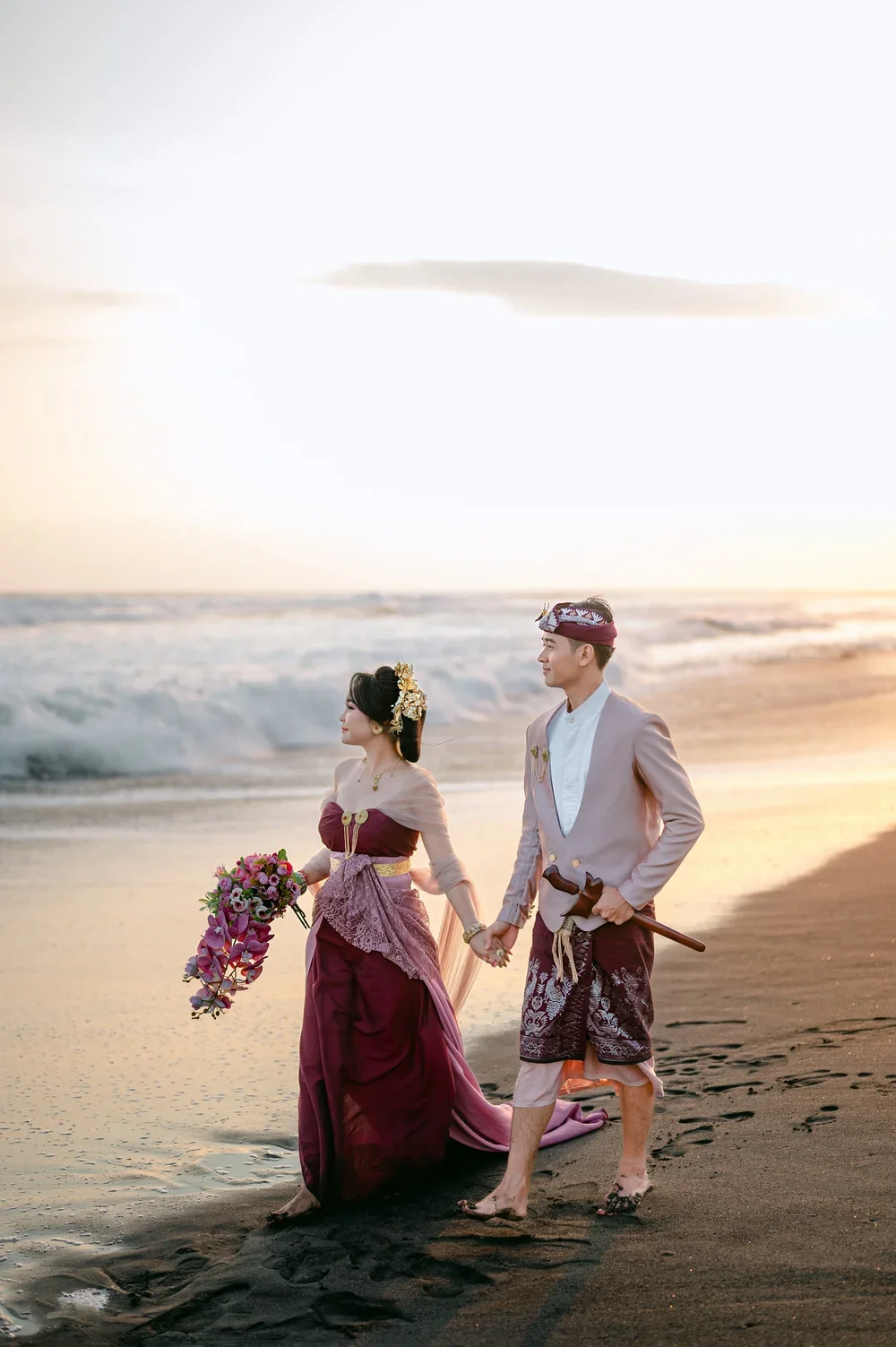 A couple dressed in traditional Thai wedding attire walking hand-in-hand along the beach during sunset, with the bride holding a bouquet of flowers and the groom carrying a sword.