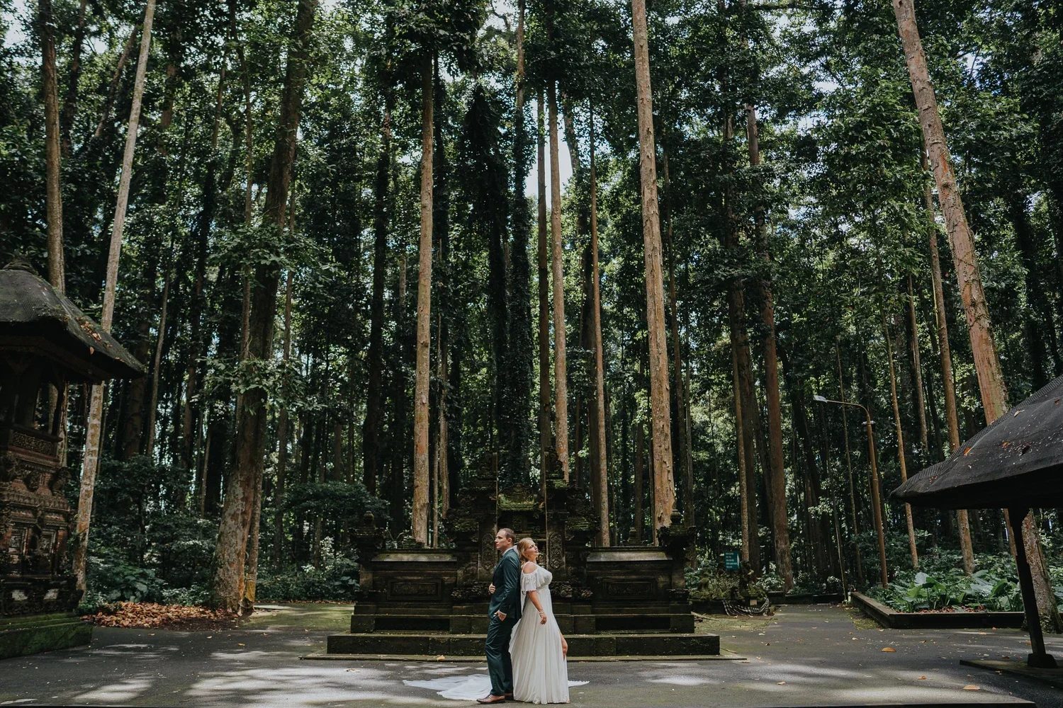 A bride and groom standing back to back outdoors in a forest with tall trees surrounding them and an altar in the background.