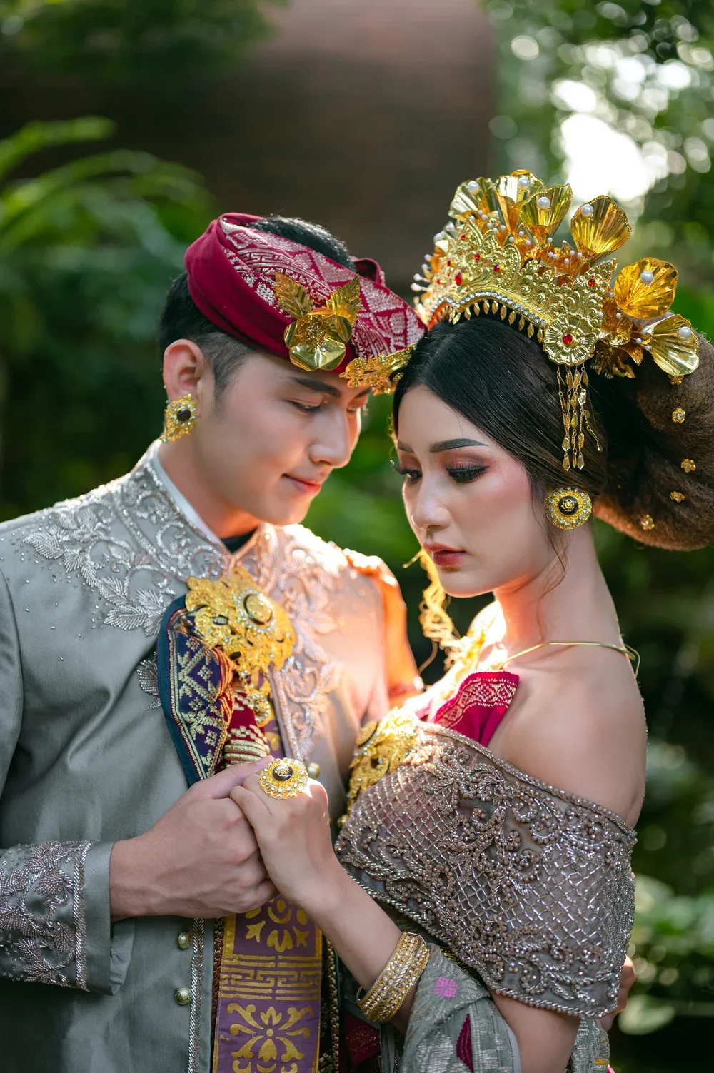 A couple dressed in traditional Indonesian wedding attire, standing close and holding hands, with a blurred green foliage background.