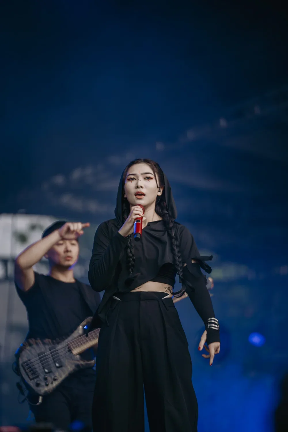Female singer performing on stage with a guitarist in the background, both dressed in black, under dark lighting.