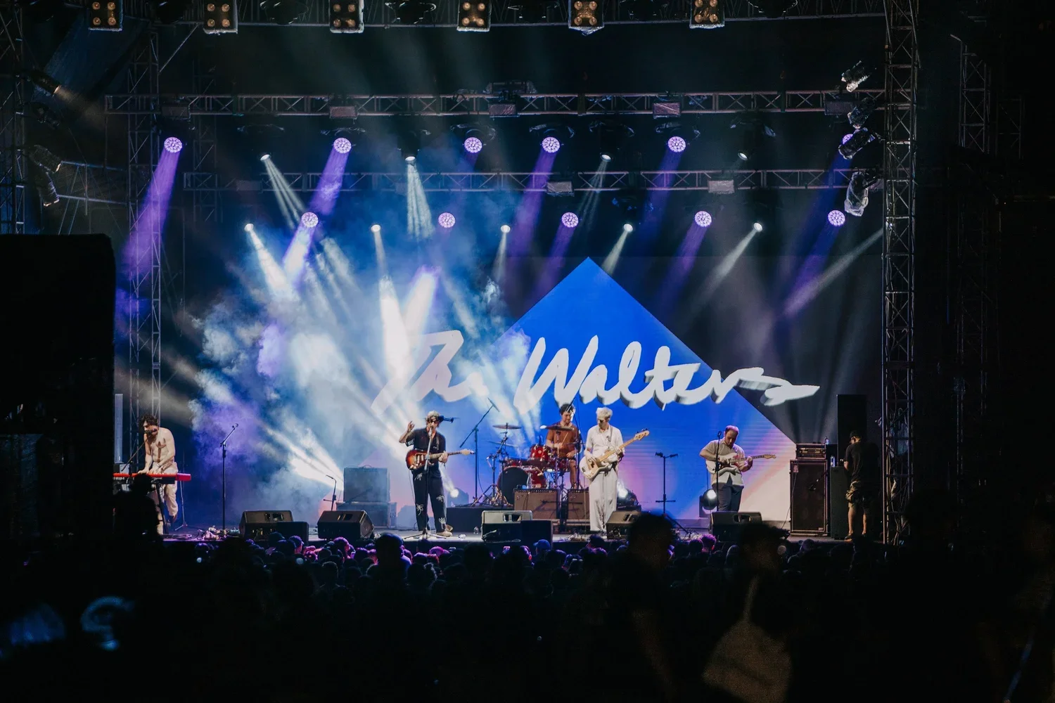 A band performing live on stage with purple and white lights, a large sign reading 'Z. Walters' in the background, and a crowd watching in front.