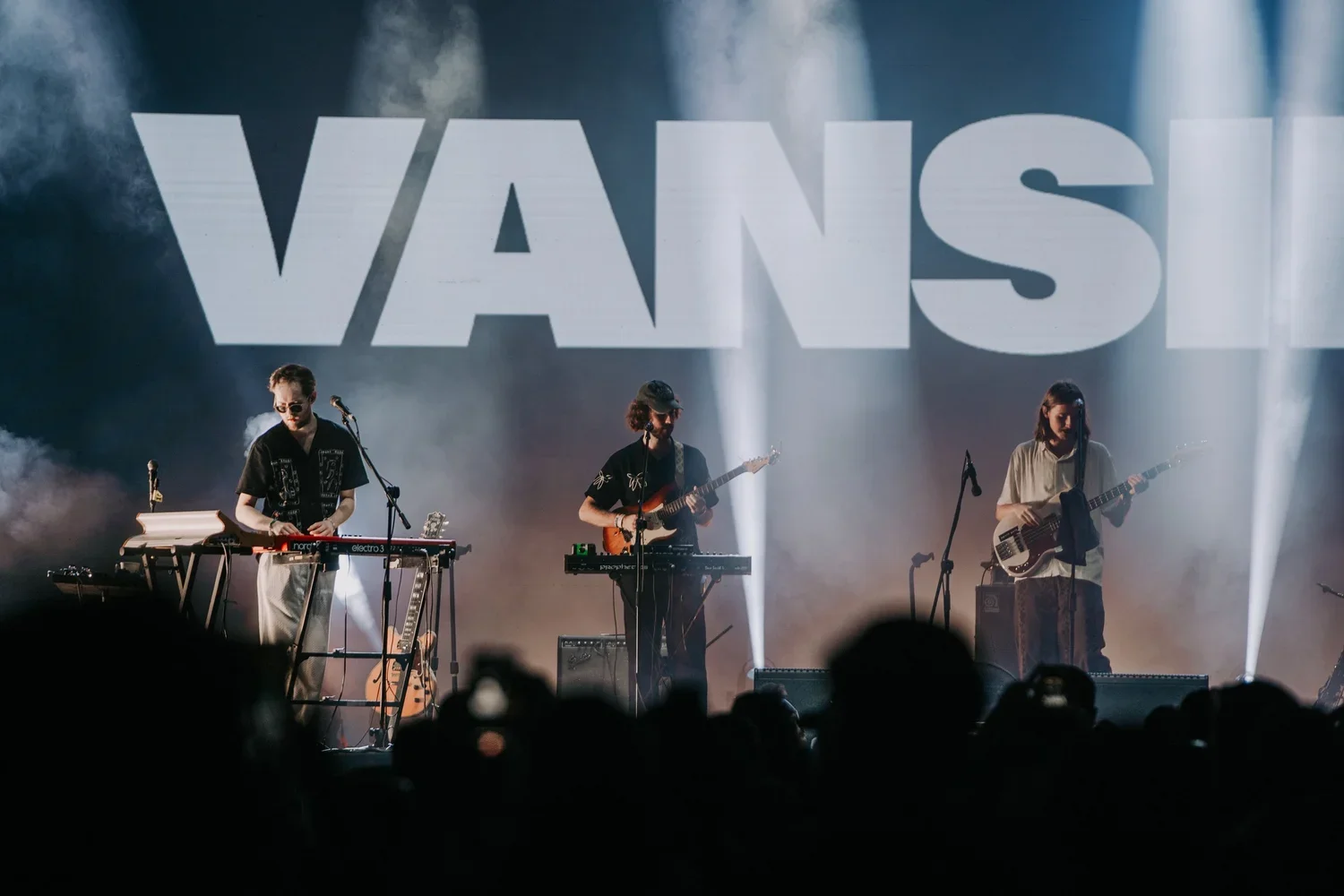 A band of three musicians performing on stage with a large 'VANSL' sign behind them, playing instruments including a keyboard, guitar, and bass, with stage lights and fog effects.