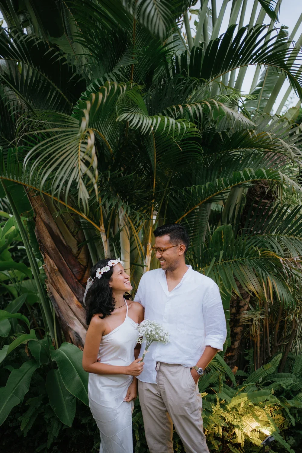 A smiling woman with a flower crown holding a bouquet of white flowers, standing next to a man with glasses, both dressed in white, in front of lush tropical greenery.