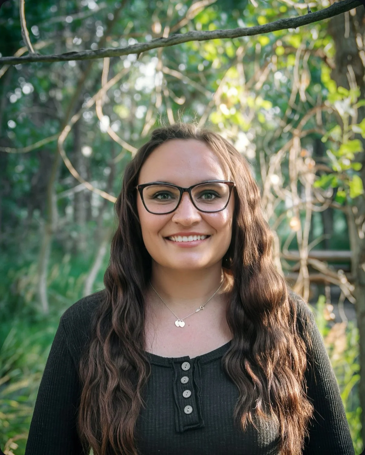 Person with long brown hair, wearing glasses and a white sweater, smiling in a studio portrait against a light background.