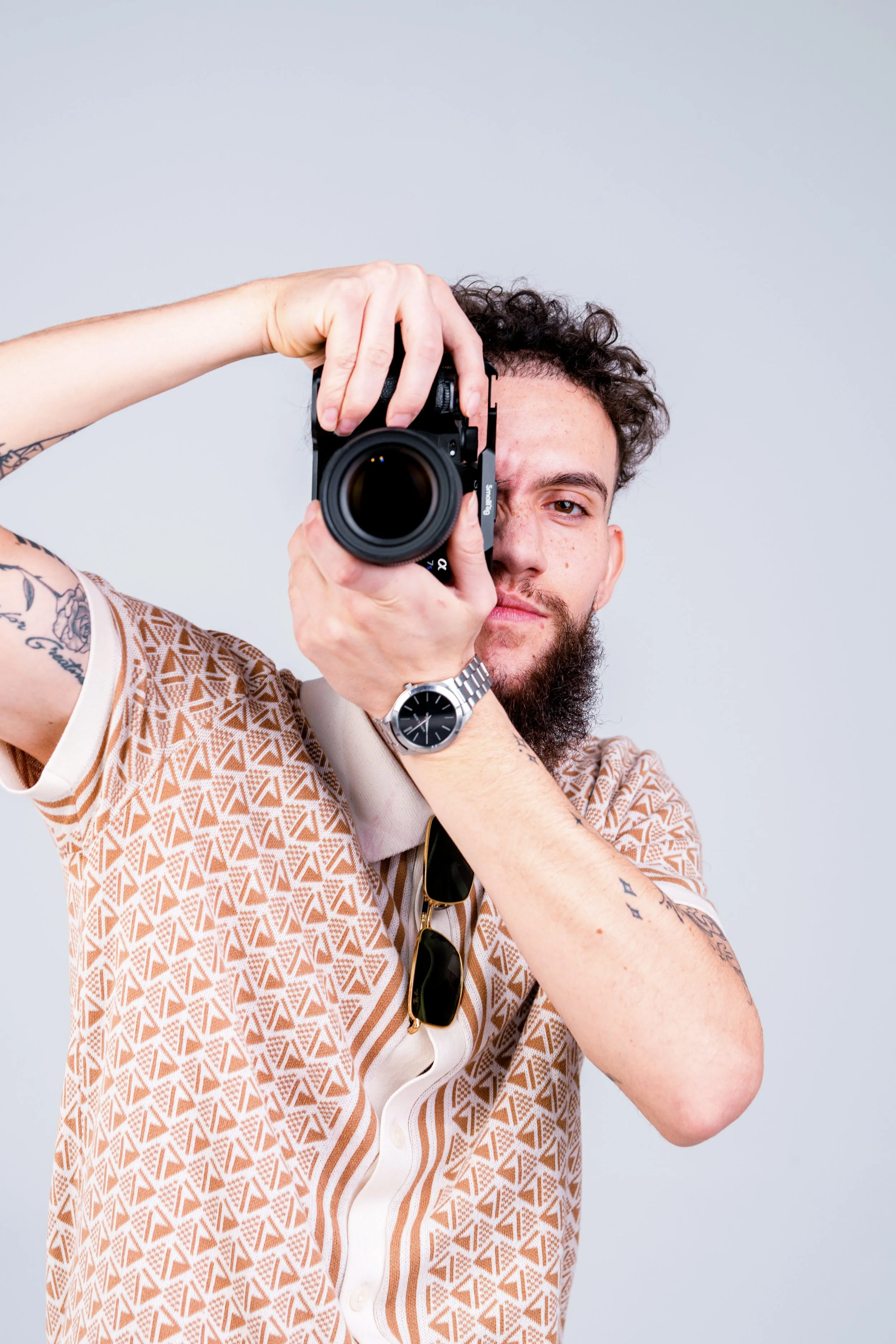 Person holding a camera, wearing a patterned shirt and wristwatch, posing for a photo.