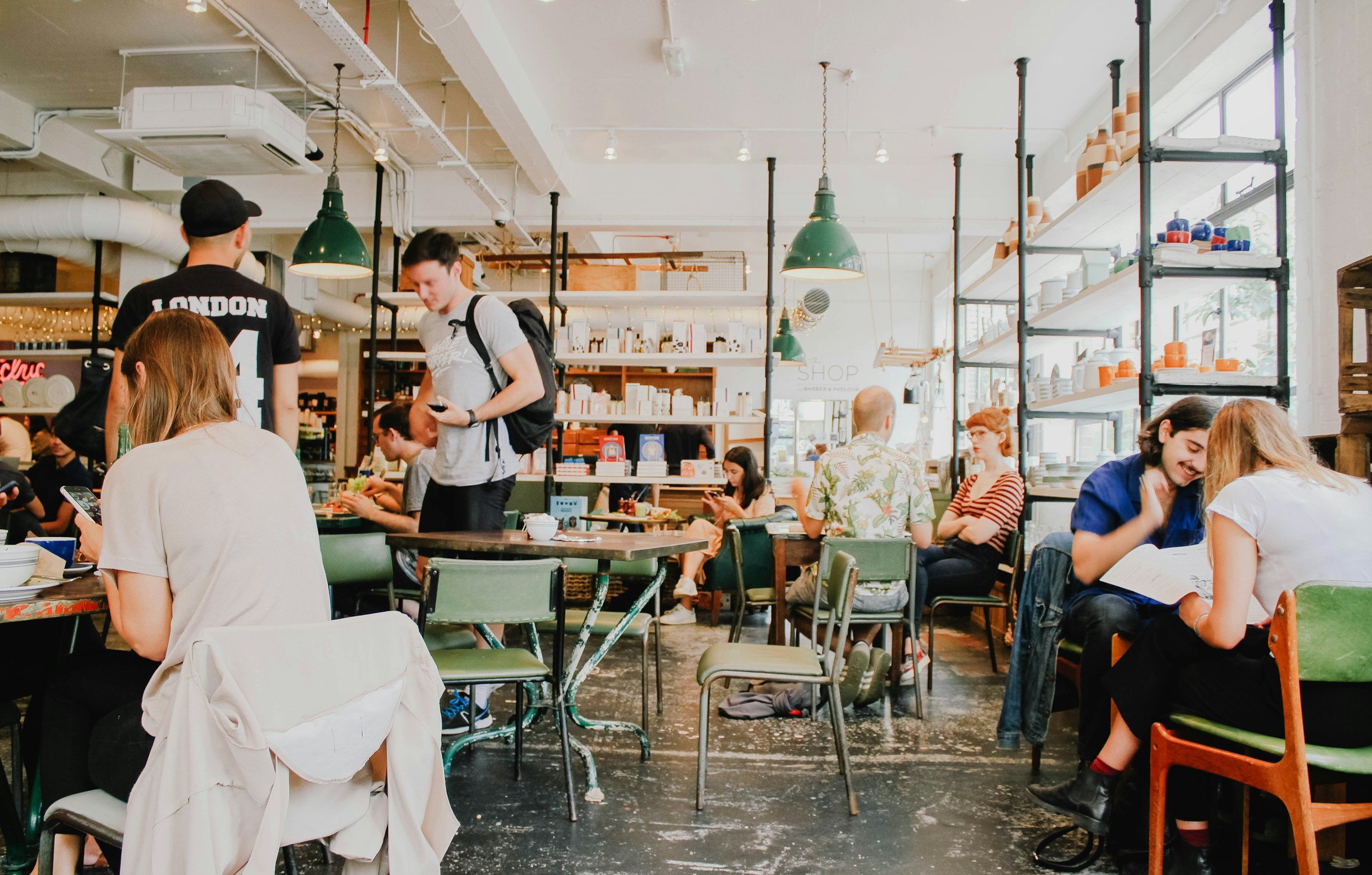 People sitting and standing inside a modern coffee shop with industrial decor, green pendant lights, and large windows