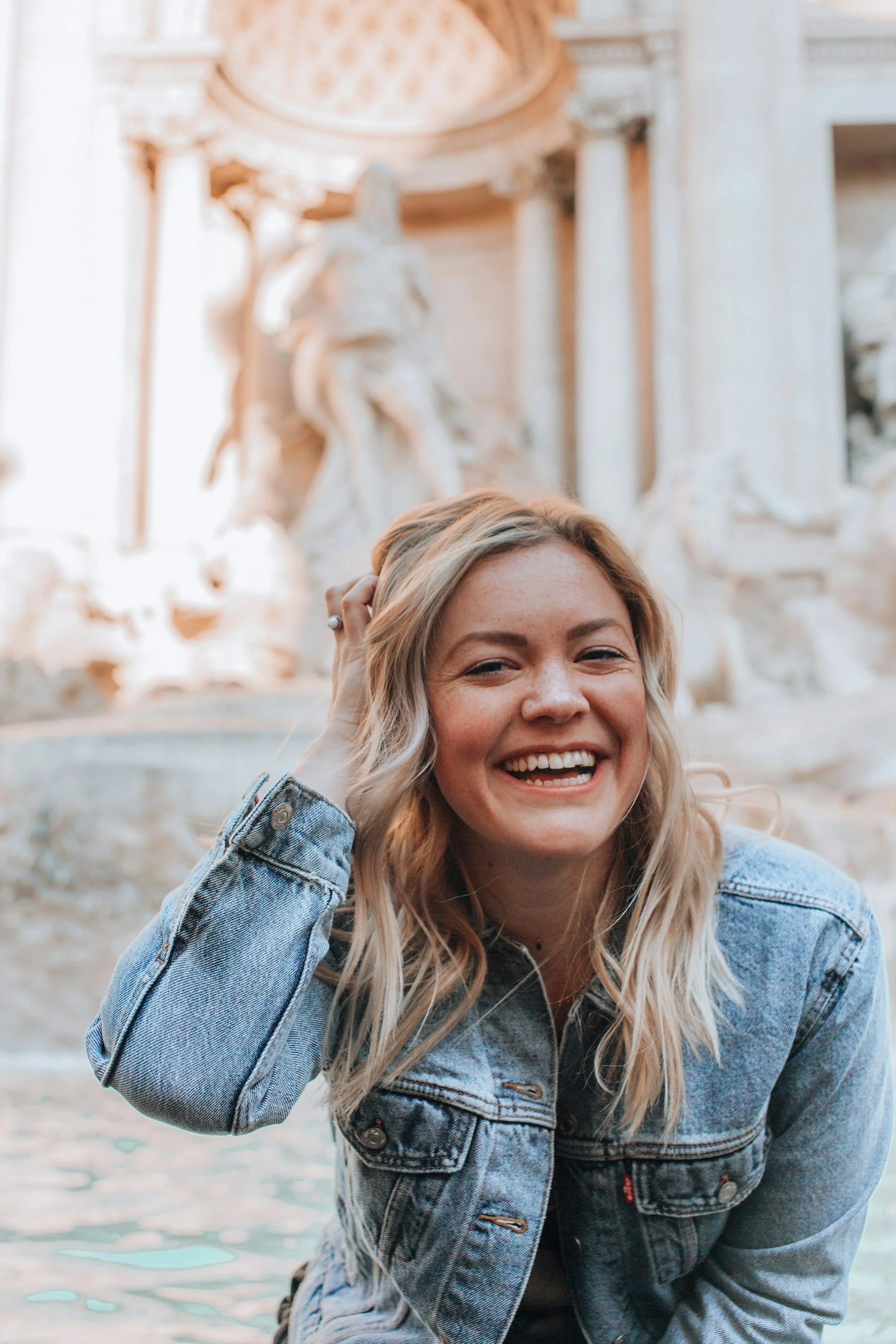 Smiling woman in a denim jacket in front of a classical fountain.