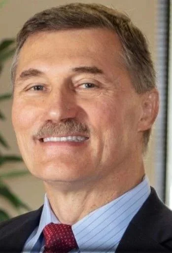 A middle-aged man with short brown hair, a mustache, wearing a suit and tie, smiling in an office environment.