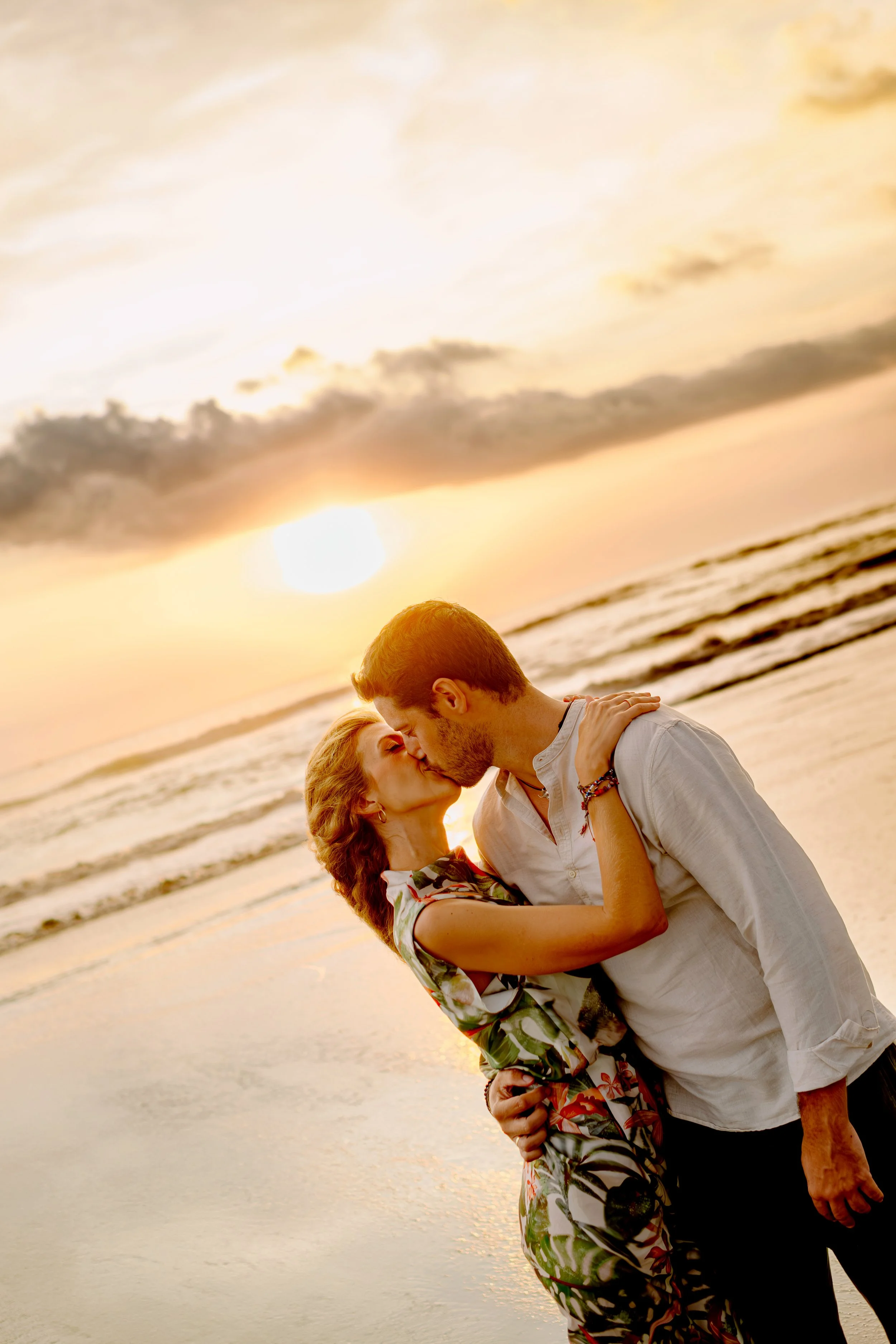 A couple kissing on the beach at sunset, with waves in the background.