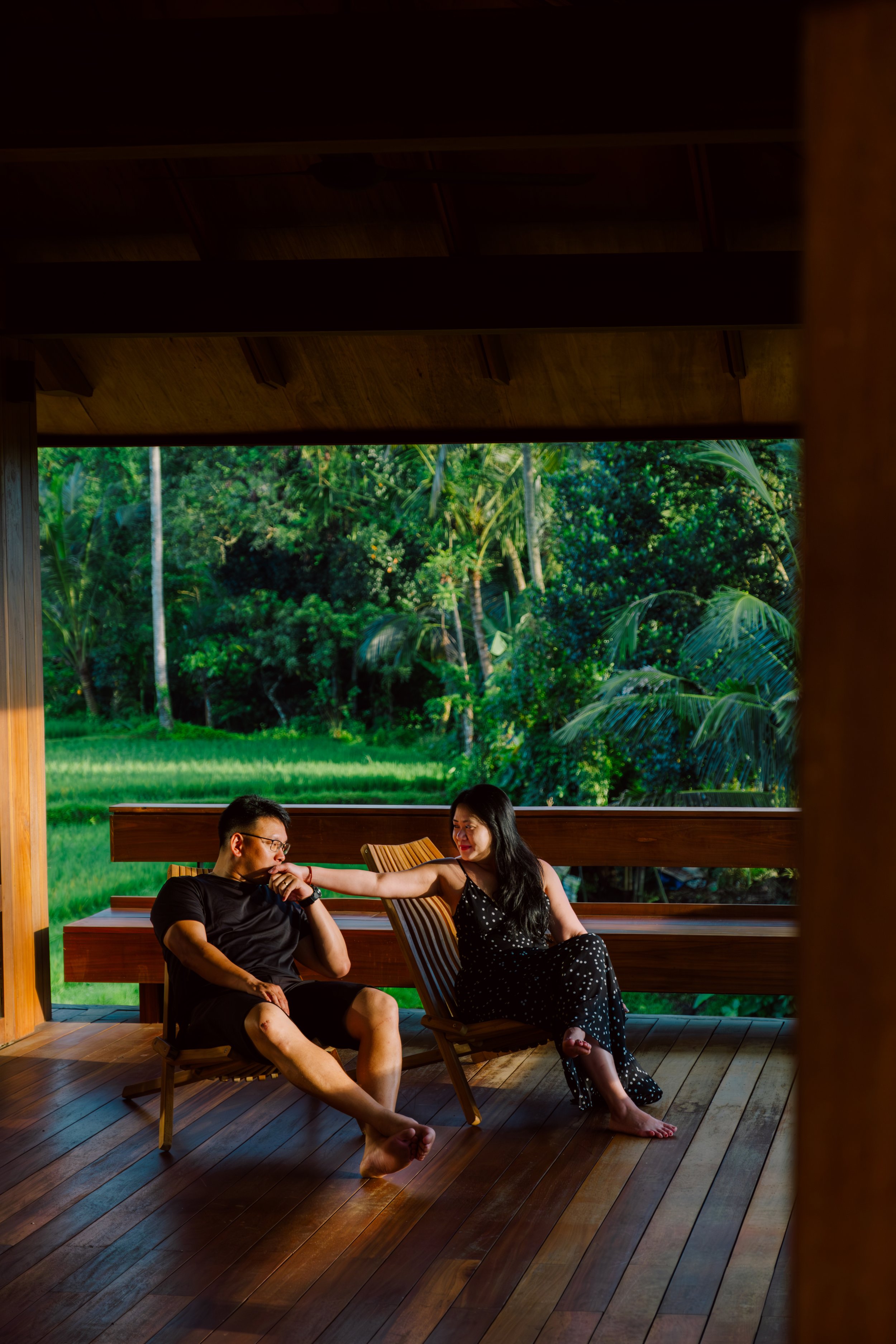 A man and woman sitting on wooden chairs on a porch overlooking lush green tropical foliage; the woman touches the man's chin.