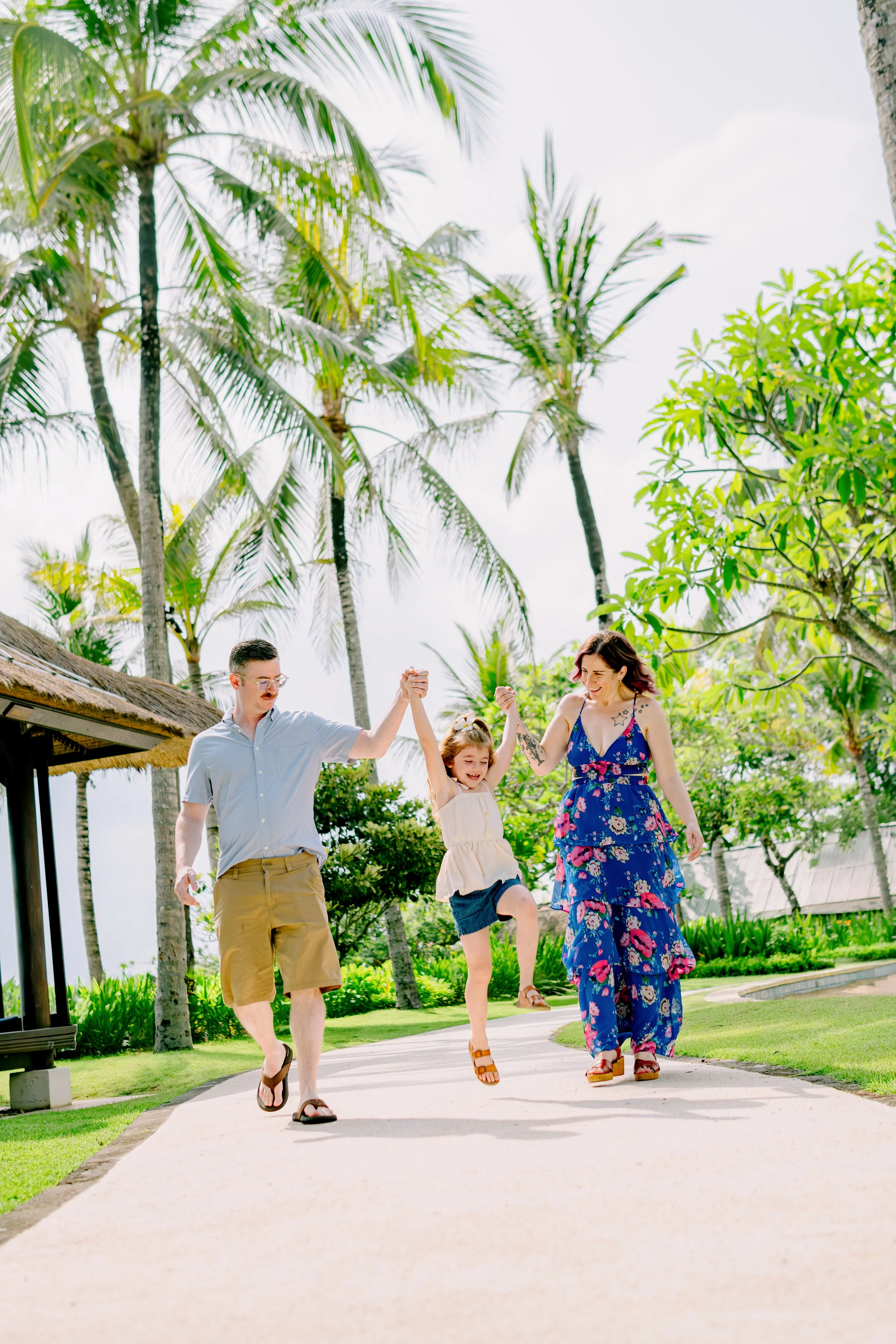 A family of three, a man, woman, and young girl, walking on a winding path in a tropical setting with tall palm trees and lush green foliage. The young girl is jumping in the air while the parents hold her hands.