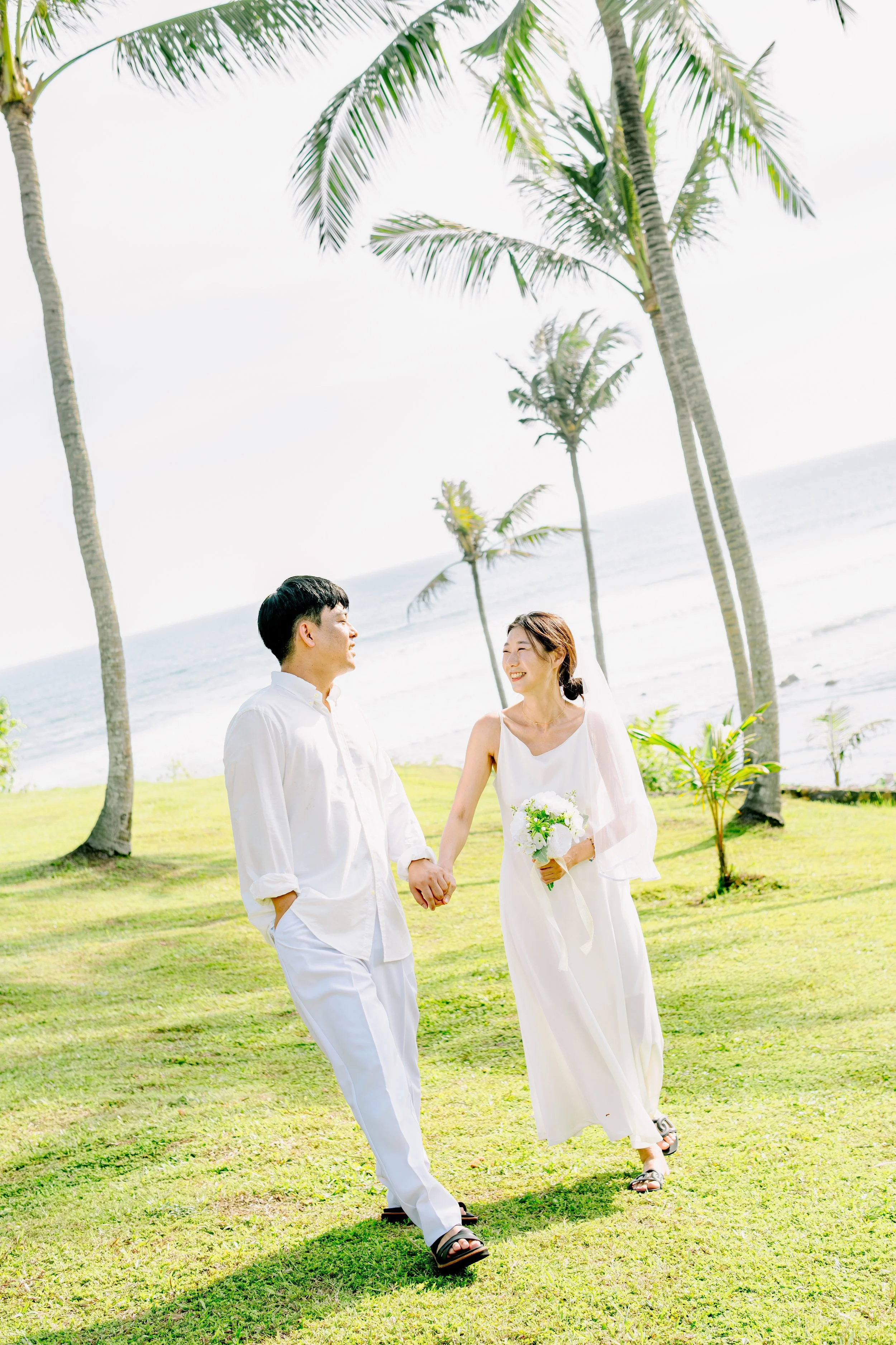 A wedding couple holding hands on a grassy tropical beach with palm trees and ocean in the background.