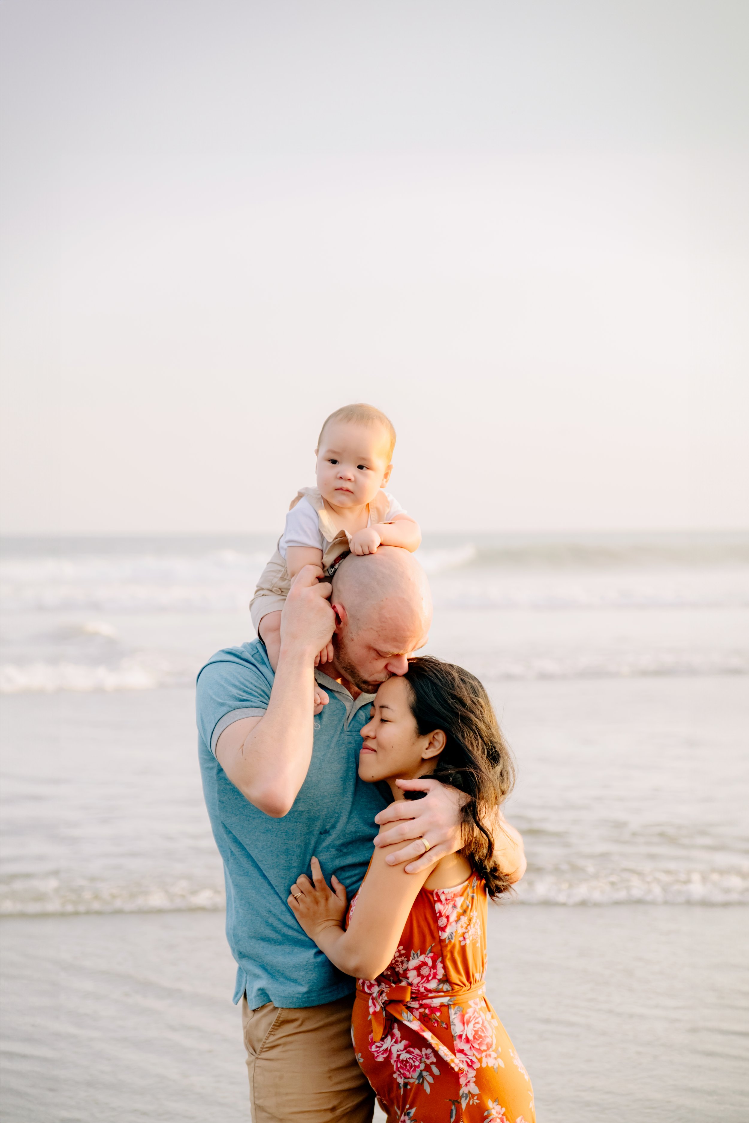 A family embracing on a beach at sunset, with a father holding a young child on his shoulders, and a mother hugging him.