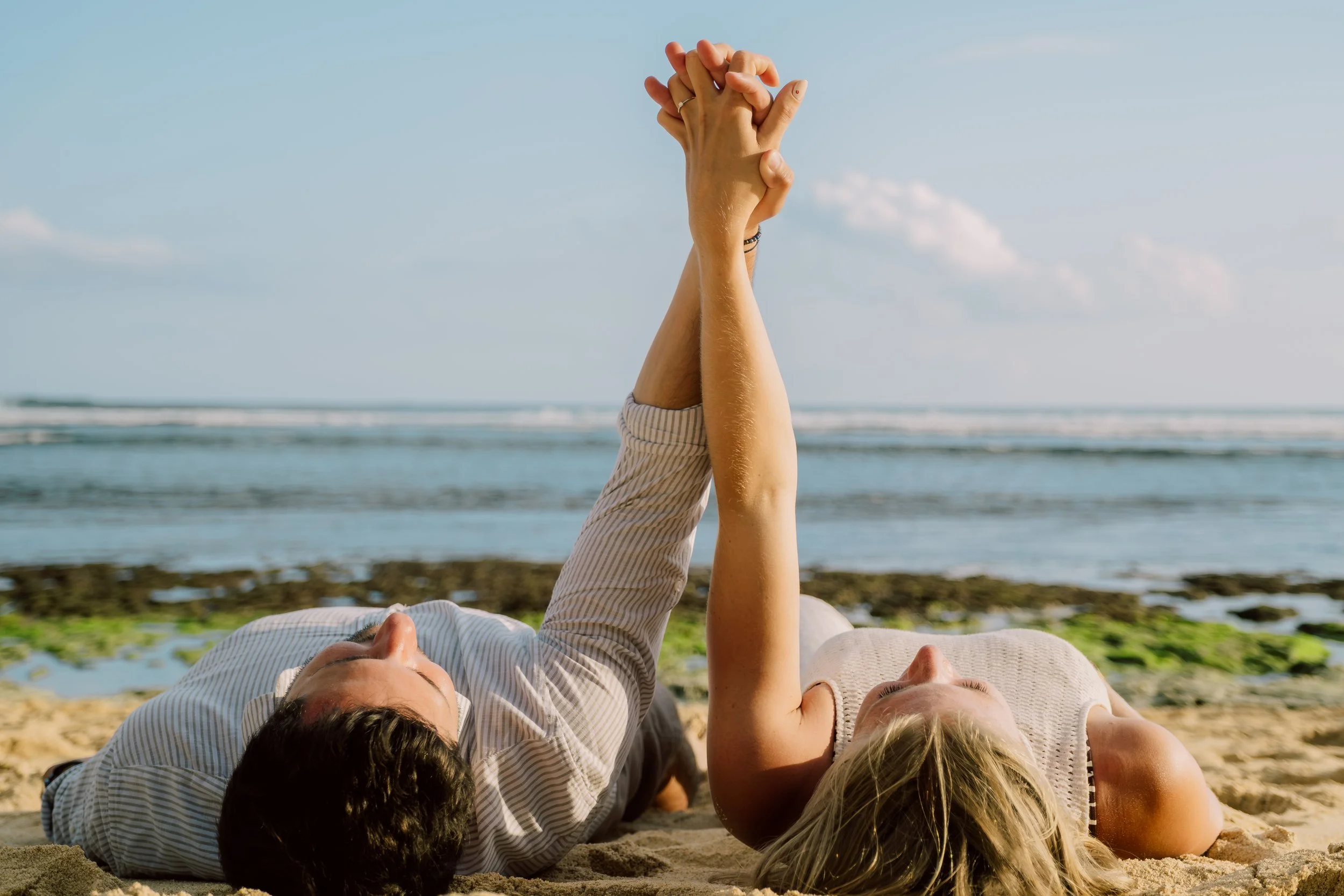 A couple lying on the beach, holding hands with their arms extended upward.