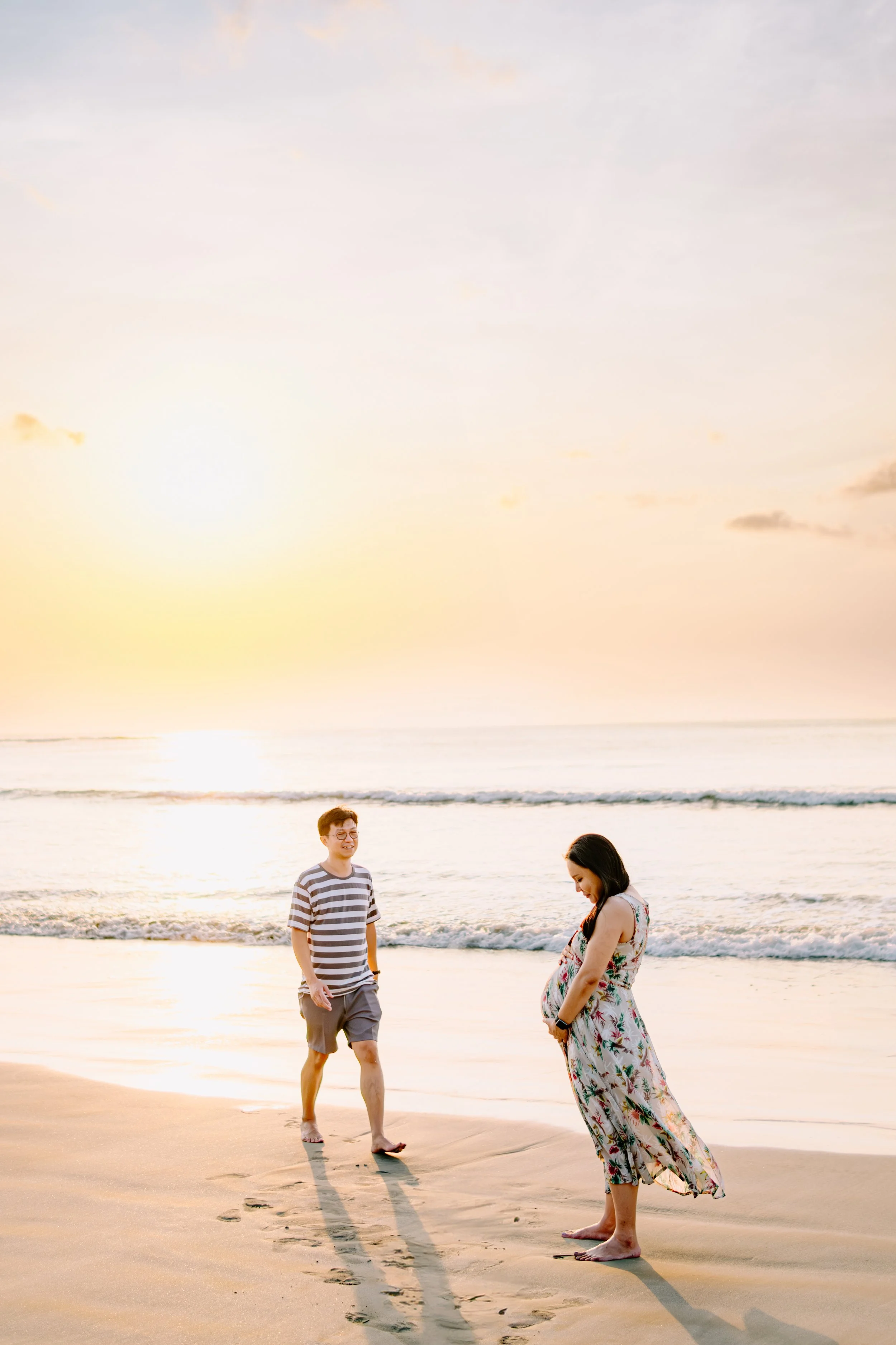 A couple on the beach at sunset, with the man smiling and the pregnant woman looking down at her belly.