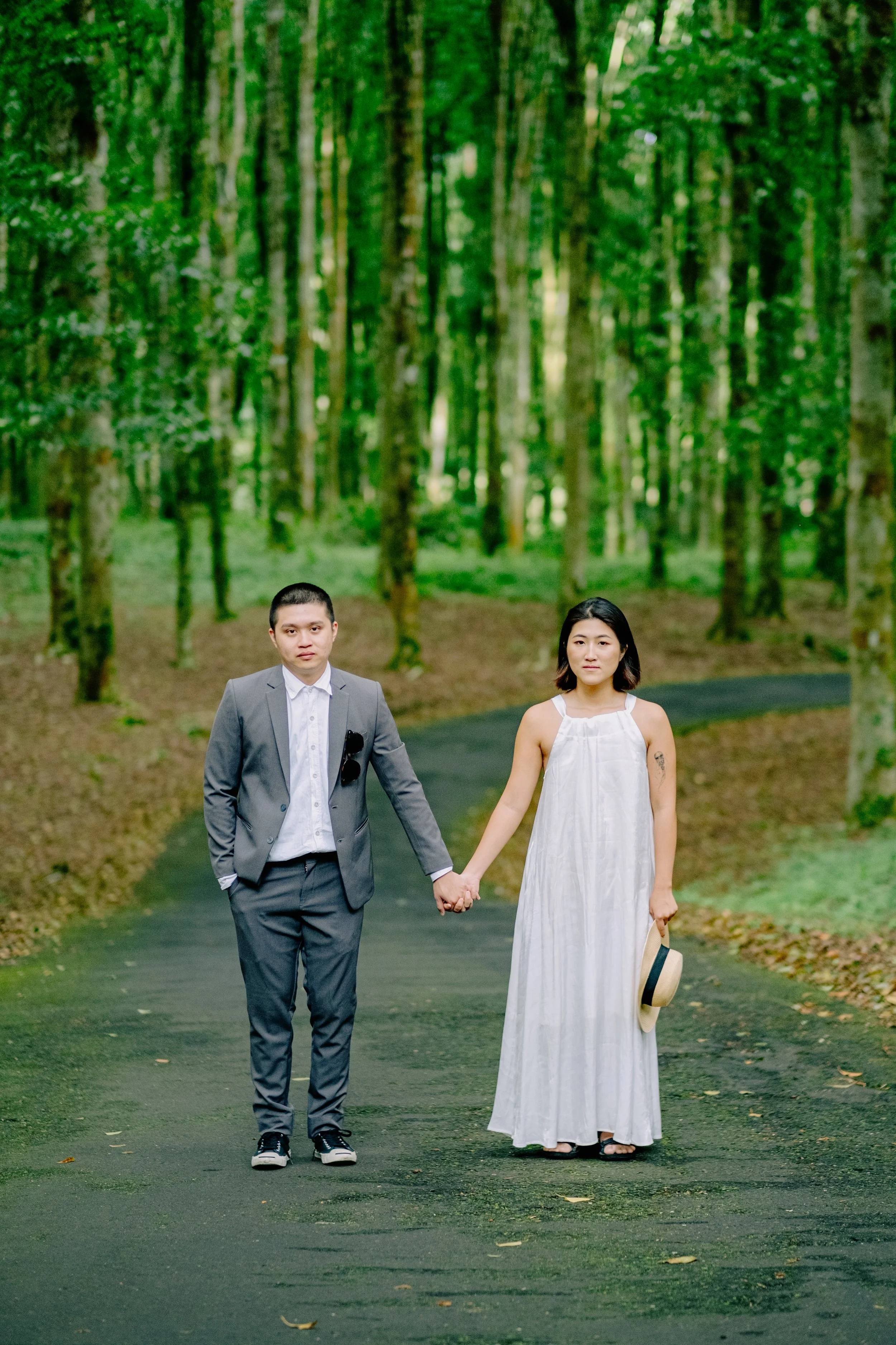 A couple holding hands walking on a forest path, surrounded by tall green trees, woman in a white dress holding a hat, man in a gray suit.