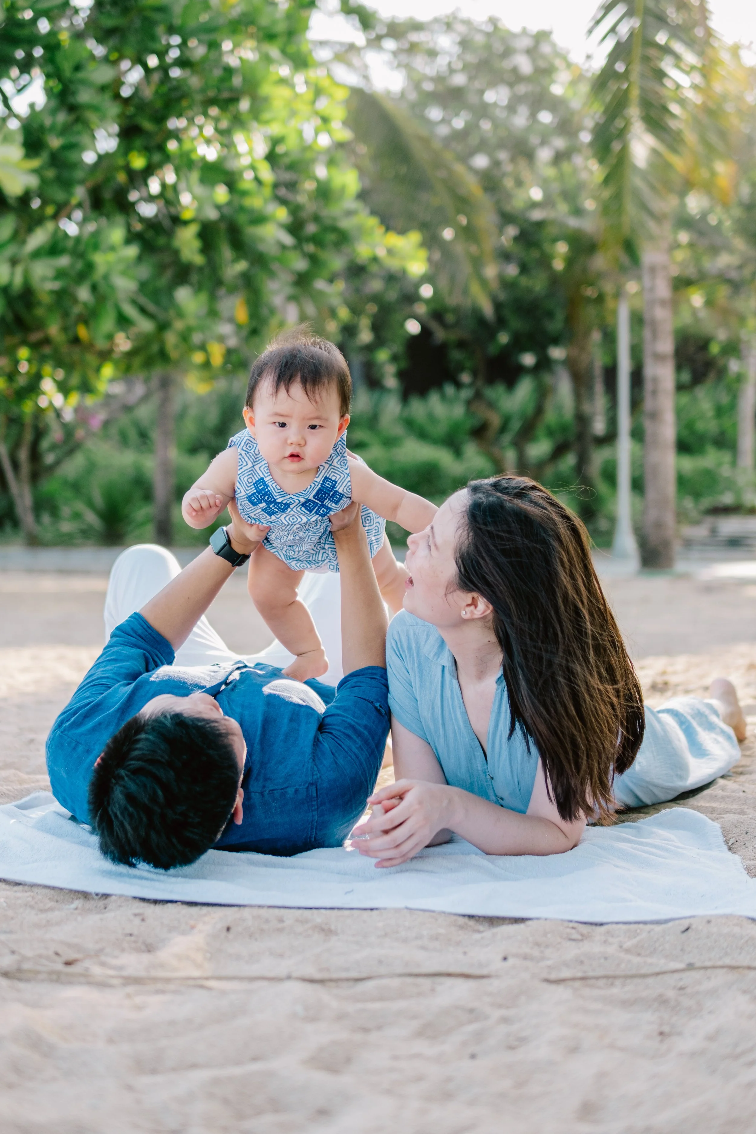 A family of three, including a man, woman, and baby, is playing on a blanket outdoors in a park or beach area with greenery and trees in the background. The man is lying on his back holding the baby in the air, while the woman watches and smiles.
