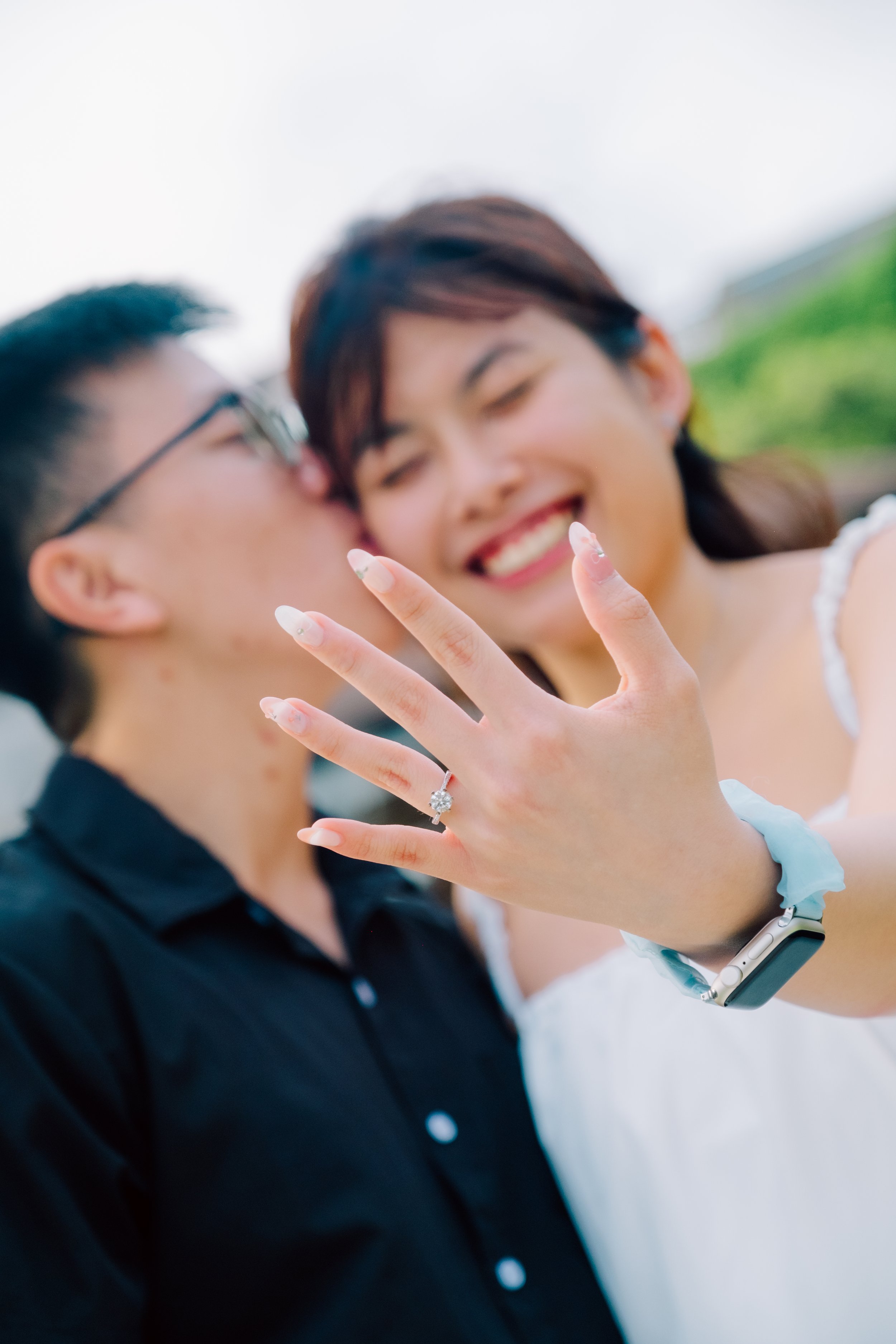 Happy woman showing off an engagement ring on her finger to a man who is kissing her on the cheek. The woman is smiling, wearing a white dress, and an Apple Watch on her wrist.