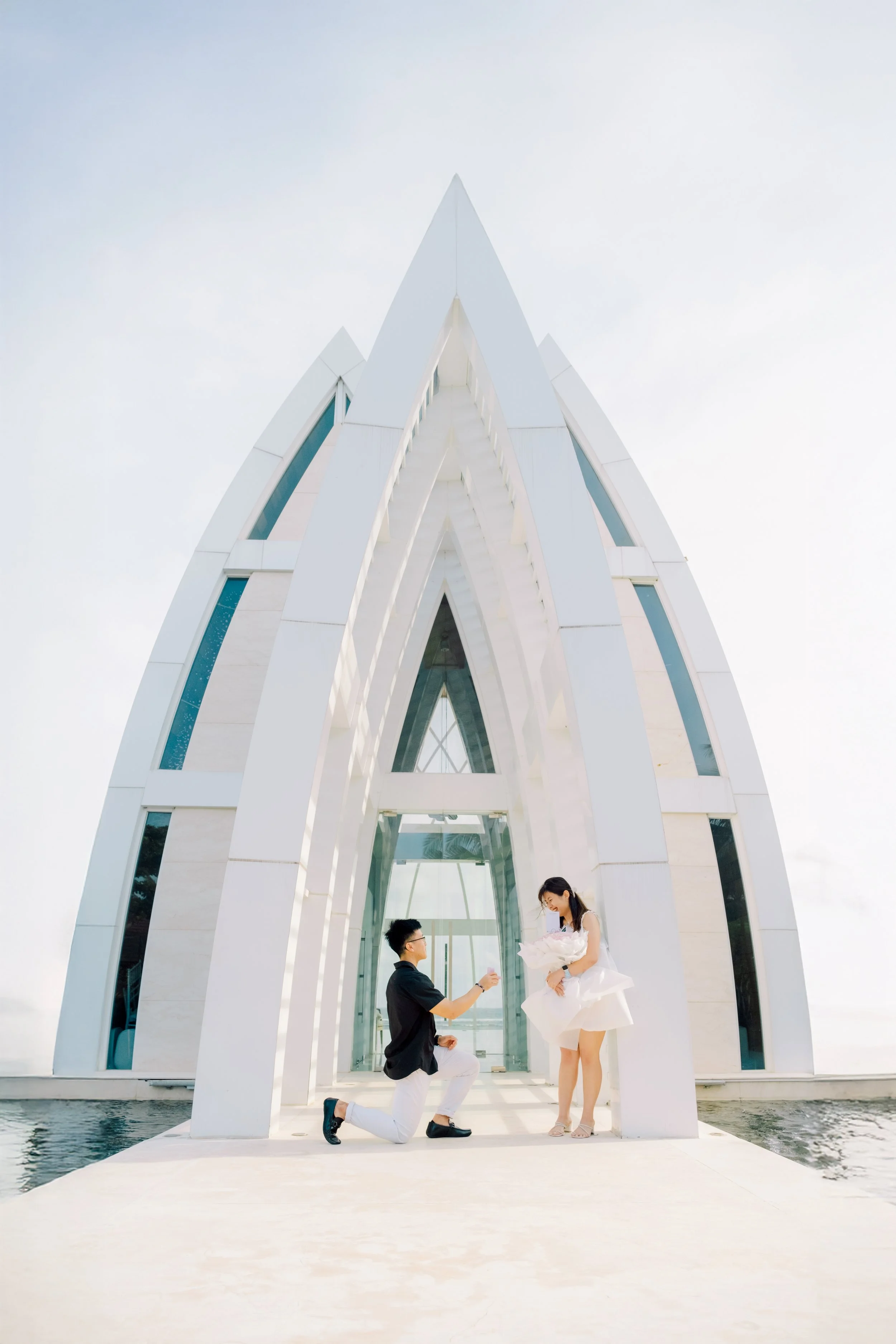 A couple at a wedding ceremony, with the man kneeling on one knee and the woman holding a bouquet in front of a modern white building with tall, pointed arches and glass windows.