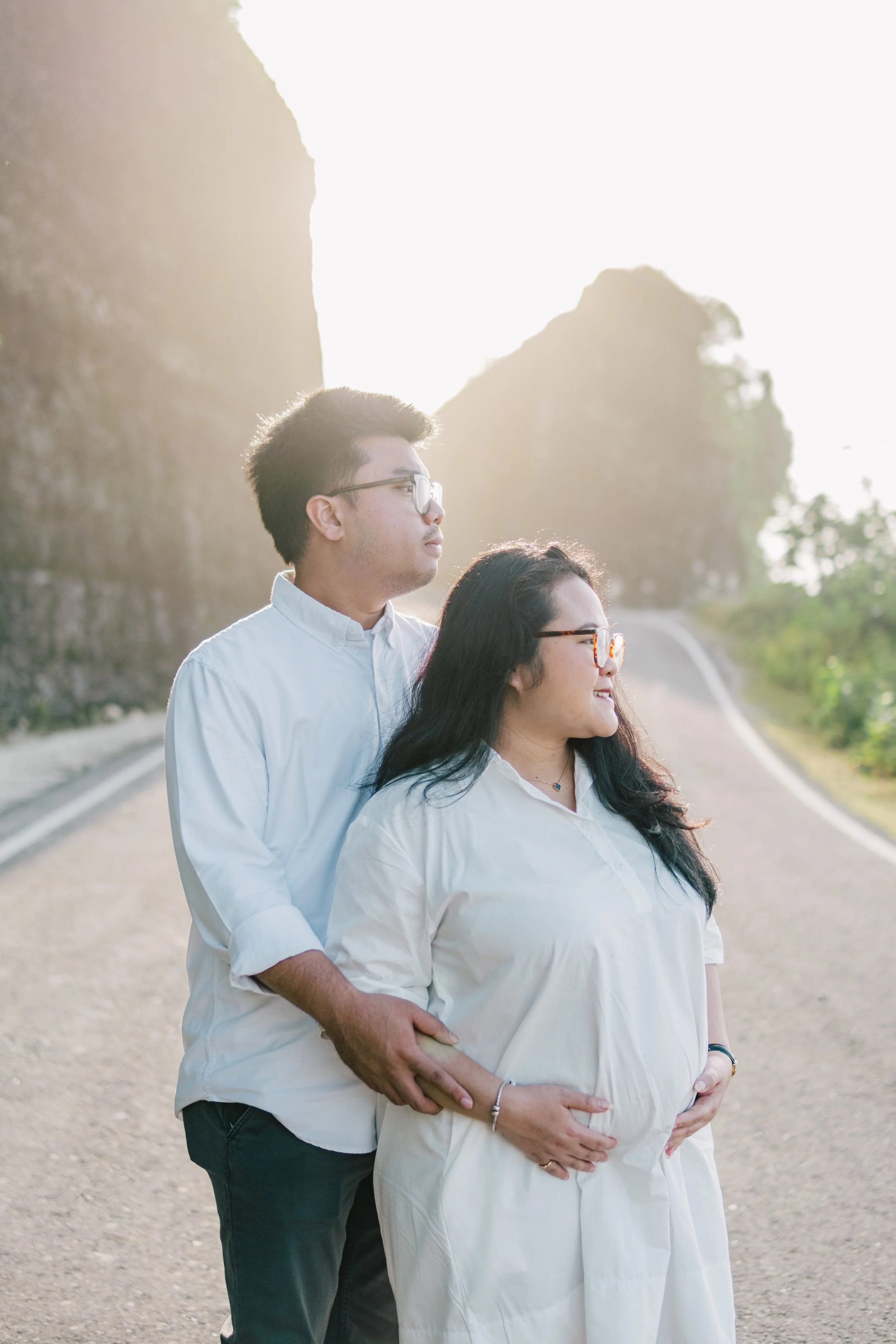A pregnant woman and a man standing on a scenic mountain road during sunrise or sunset, both wearing white shirts and glasses, with the man gently holding the woman from behind.