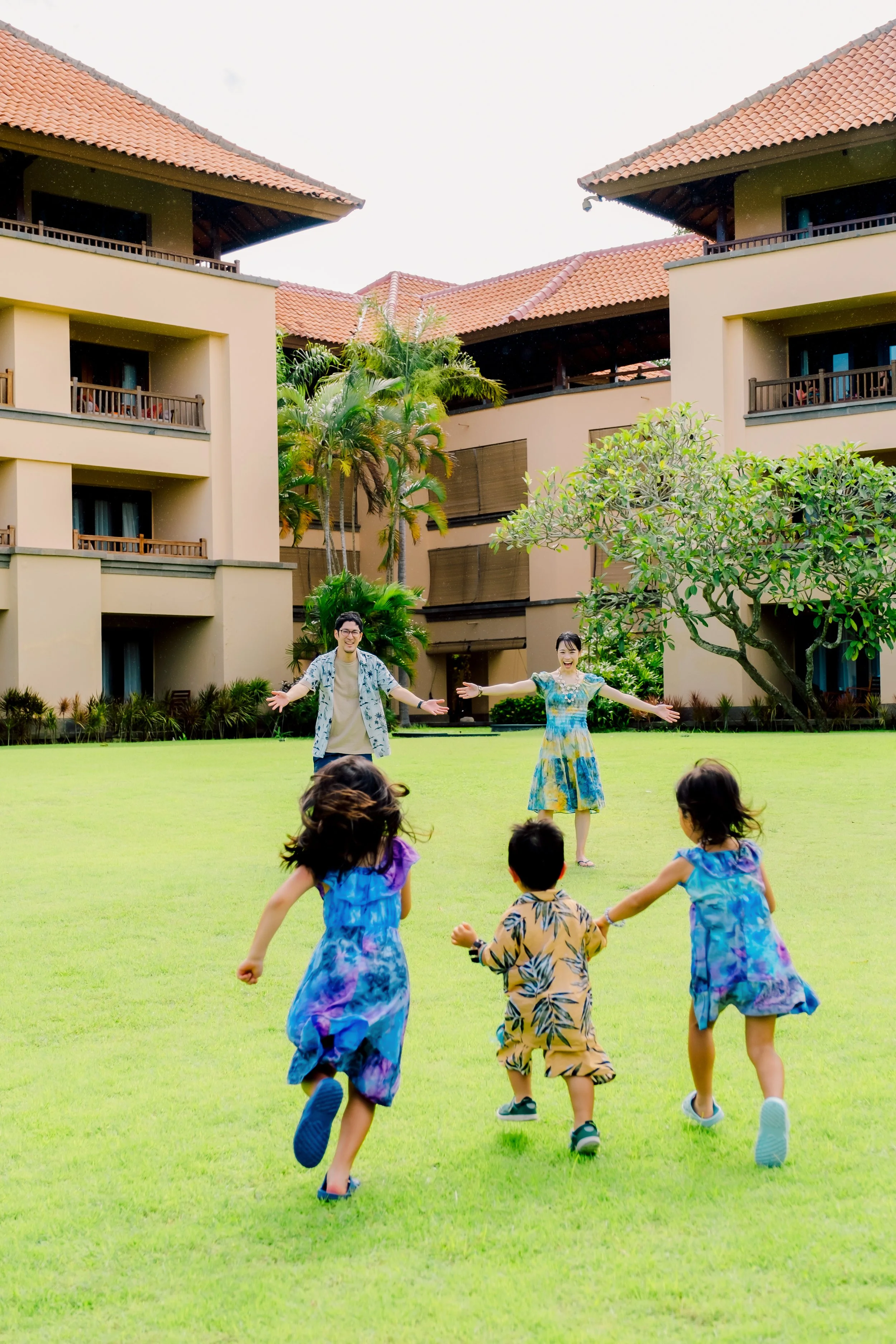 A group of children running towards two adults with arms wide open on a green lawn in front of a beige building with a red tile roof and tropical plants.
