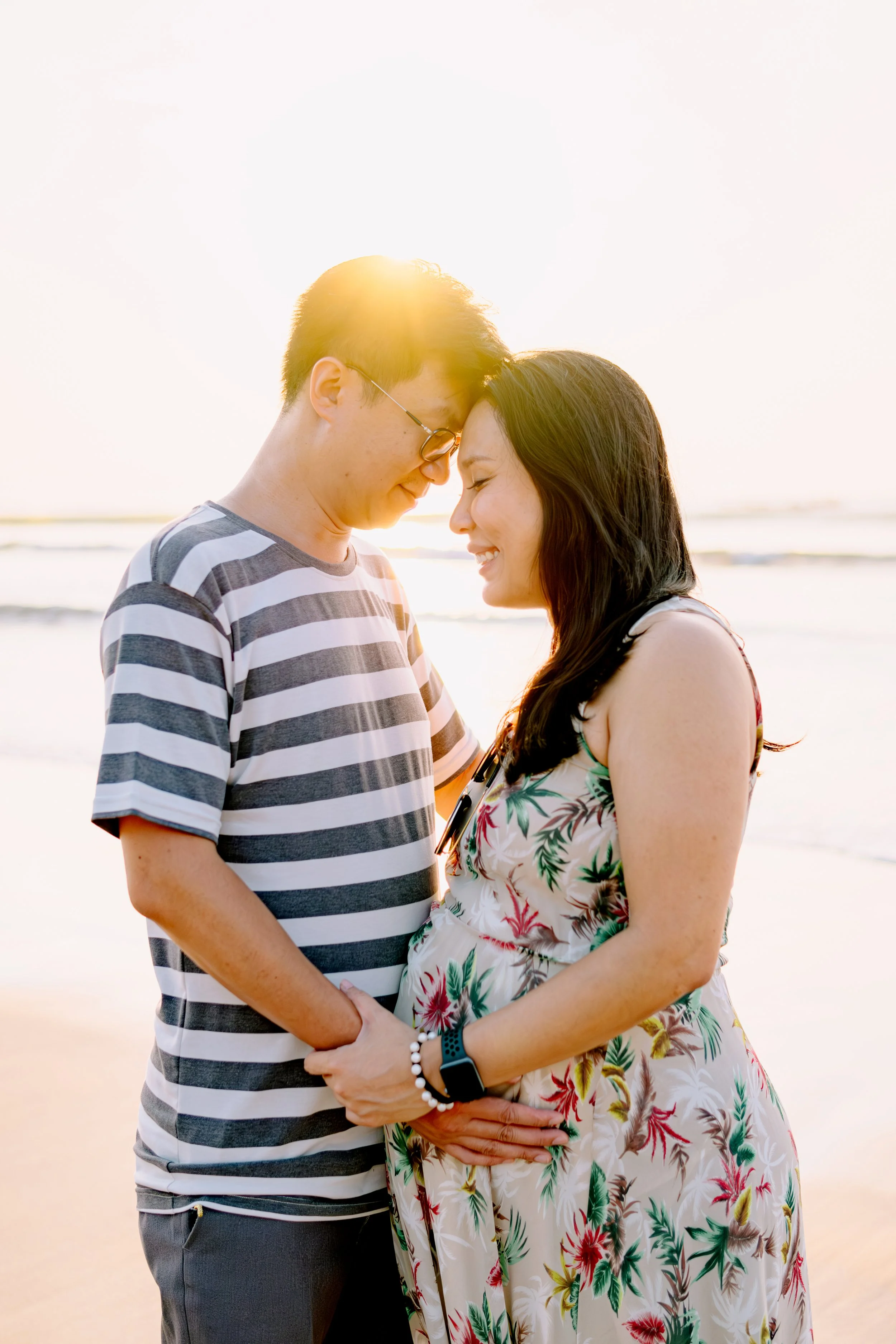 A couple standing close together on the beach at sunset, with their foreheads touching and smiling, the man wearing a striped t-shirt, and the woman in a floral dress, with the ocean waves in the background.