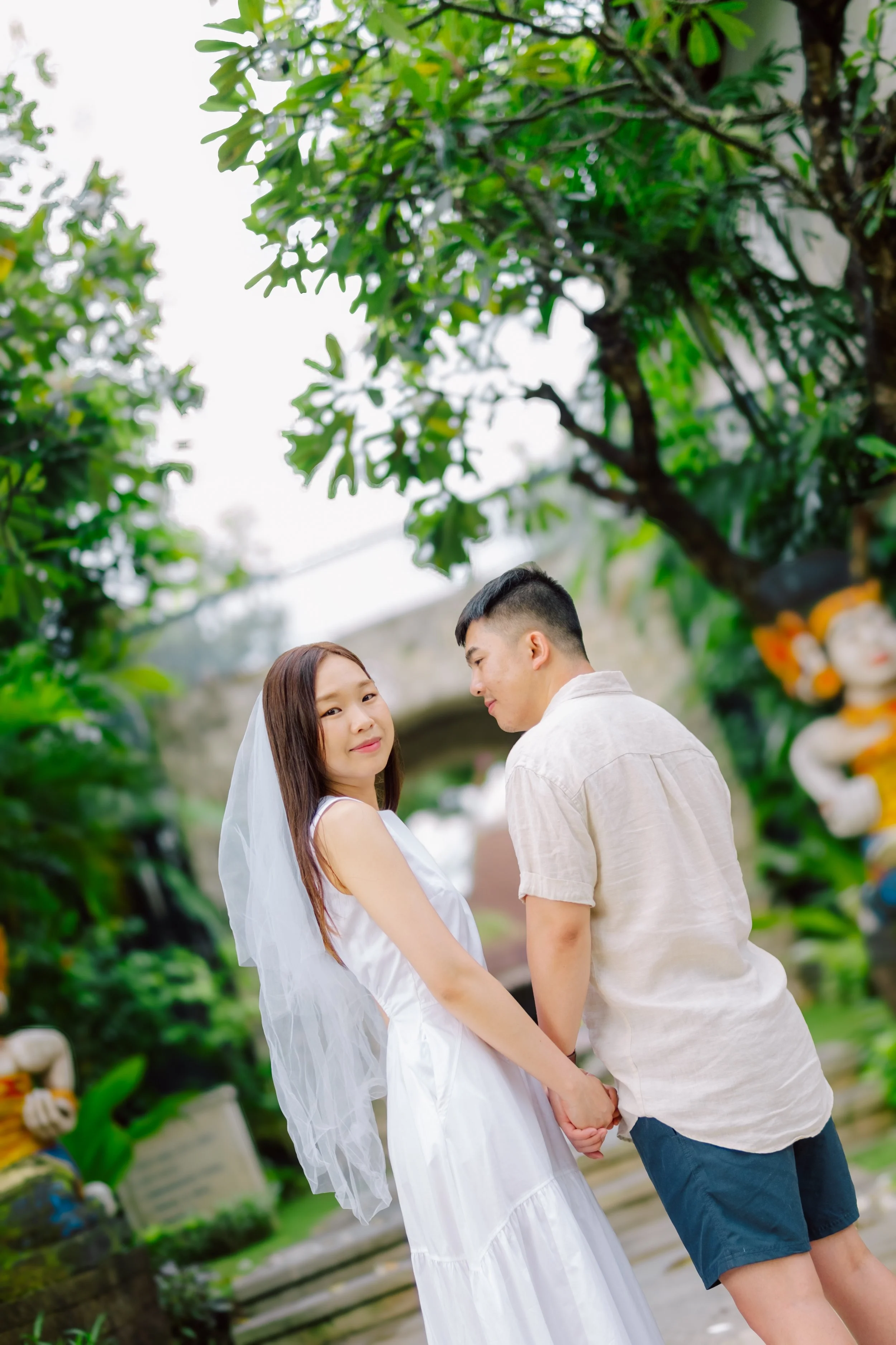 A young woman in a white dress and veil holding hands with a young man in a beige shirt and navy shorts, standing outdoors under green trees, with a stone arch and colorful statue in the background.