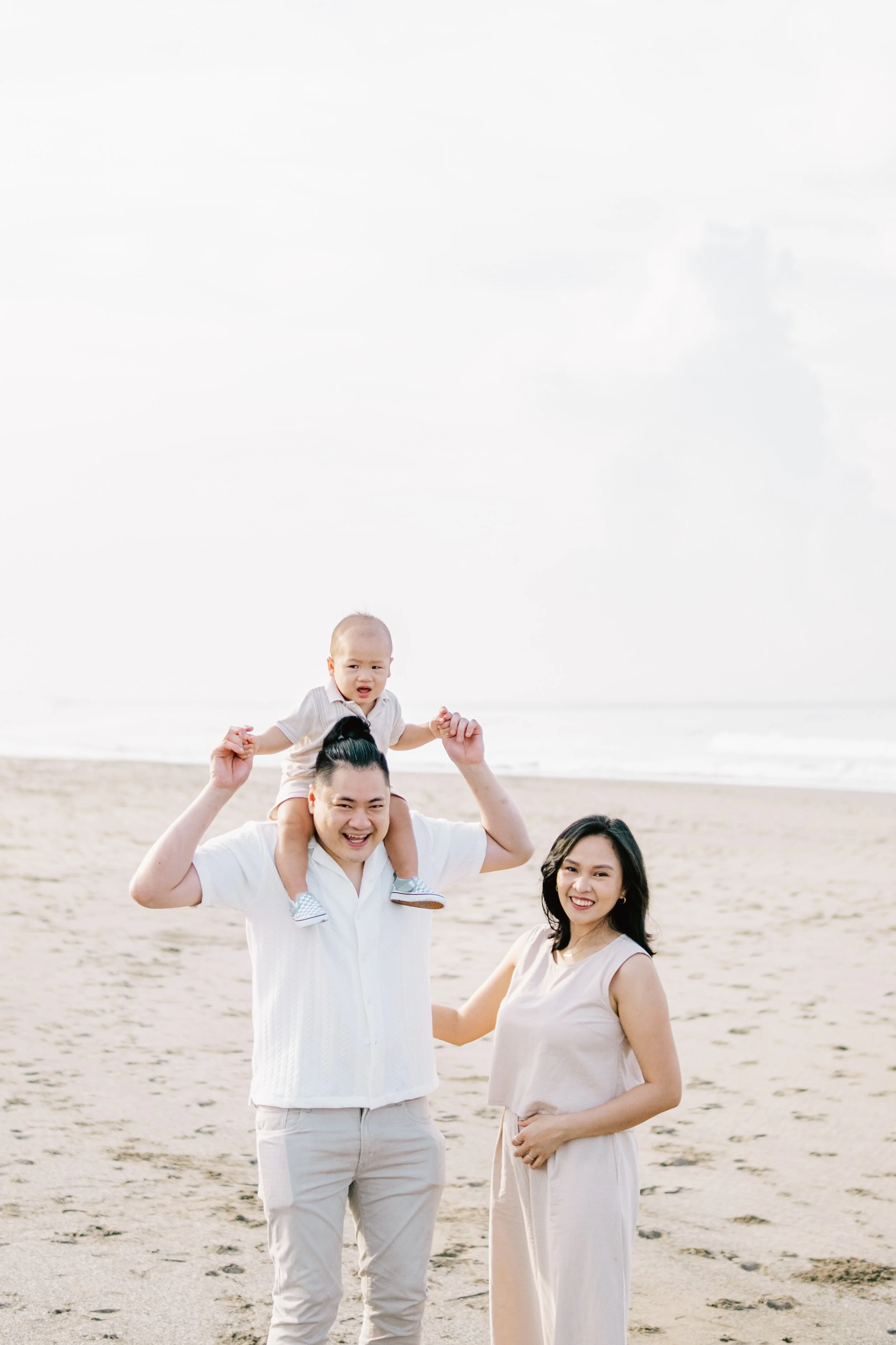 A family of three enjoying time together on the beach, with the father carrying their young child on his shoulders and the mother standing beside them.